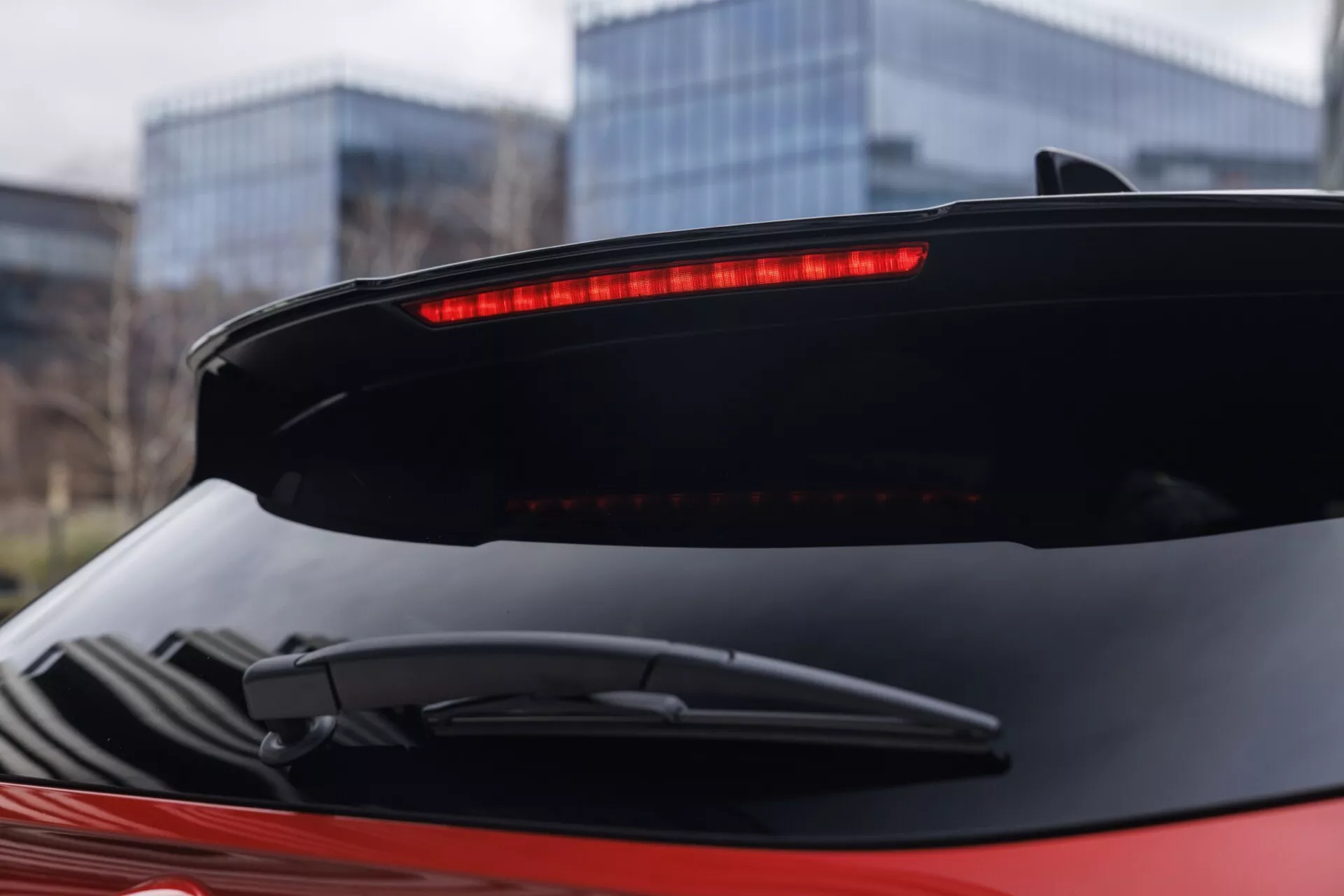 Detailní pohled na zadní část vozu Nissan Qashqai 2024 (J12-facelift) s důrazem na zadní spoiler s osvětleným vysoko připevněným LED brzdovým světlem a horizontálně parkovaným zadním stěračem. Obrázek je pořízen z mírného nízkého úhlu a předvádí aerodynamické provedení šikmého zadního skla a bezproblémovou integraci víka kufru. Červená barva karoserie kontrastuje s černým spojlerem a stěračem, na neostrém městském pozadí.
