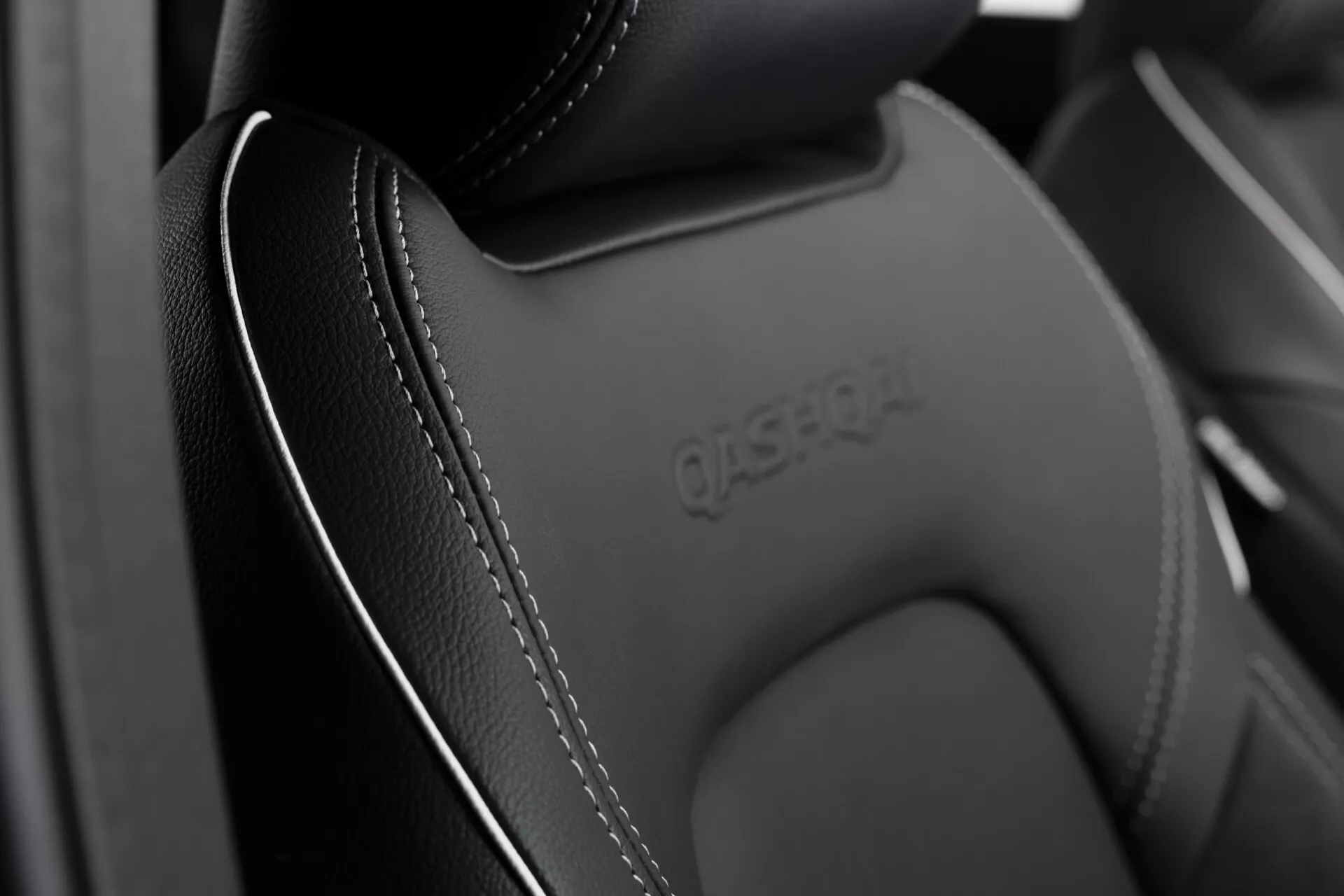 Detailní pohled na horní část autosedačky modelu Nissan Qashqai 2024 (J12 Facelift). Obrázek zdůrazňuje vysoce kvalitní kožený materiál s pečlivým světlým prošíváním, tvarovanou opěrku hlavy s reliéfním logem a matný tmavý povrch. Měkké osvětlení zachycuje jemné detaily a řemeslnou zručnost interiéru luxusního nebo prémiového vozidla.