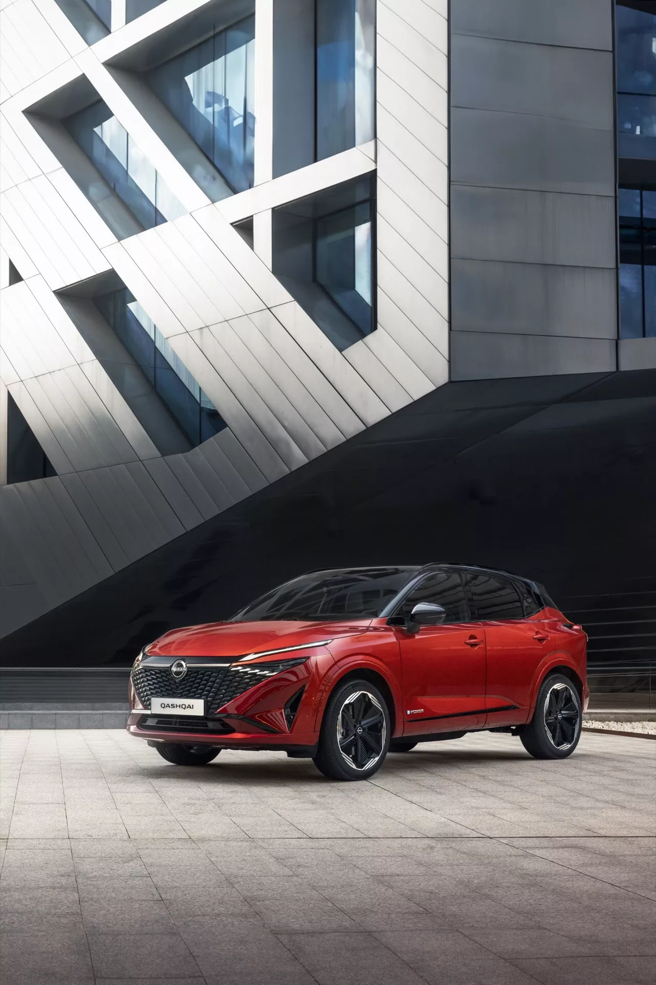 Červené SUV Nissan Qashqai 2024 (J12-Facelift) zaparkované před moderní budovou s geometrickými tvary, fotografované z nízkého, předního levého úhlu, který zdůrazňuje propracovanou přední masku, ostré LED světlomety a sportovní nárazník. Ukazuje přední čtvrt pohled včetně kapoty, dveří řidiče a jedinečných slitinových kol s dominantním postojem a futuristickým pozadím.