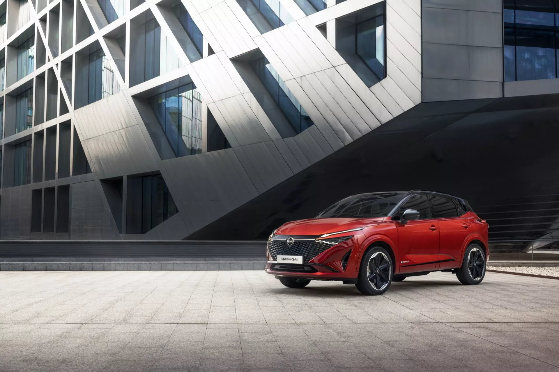 Obrázek červeného Nissan Qashqai 2024 (J12 facelift) SUV zaparkovaného na dlážděné ploše před moderní, hranatou budovou. Fotografováno z přední tříčtvrteční perspektivy, snímek zvýrazňuje výraznou texturovanou přední masku, elegantní LED světlomety, aerodynamickou tvarovanou kapotu, robustní plastové obložení kolem oblouků kol, vícepaprsková litá kola, zpětná zrcátka s integrovanými směrovkami, střešní nosiče, panoramatické střešní okno a sportovní zadní design se strmým zadním oknem a střešním spoilerem. Moderní pozadí doplňuje design SUV, což naznačuje jeho městskou univerzálnost a pokročilé funkce.