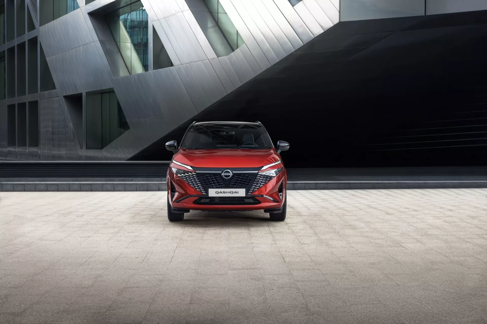 Pohled zepředu na Nissan Qashqai 2024 (J12-Facelift) s úzkými úhlovými LED světlomety, charakteristickou V-tvarovanou maskou chladiče s logem výrobce, dynamickým předním nárazníkem s možnými pokročilými senzory a jemně tvarovaným kapotou. Živá červená barva vozu kontrastuje s tlumeným pozadím moderní budovy, což zdůrazňuje jeho současný městský design.