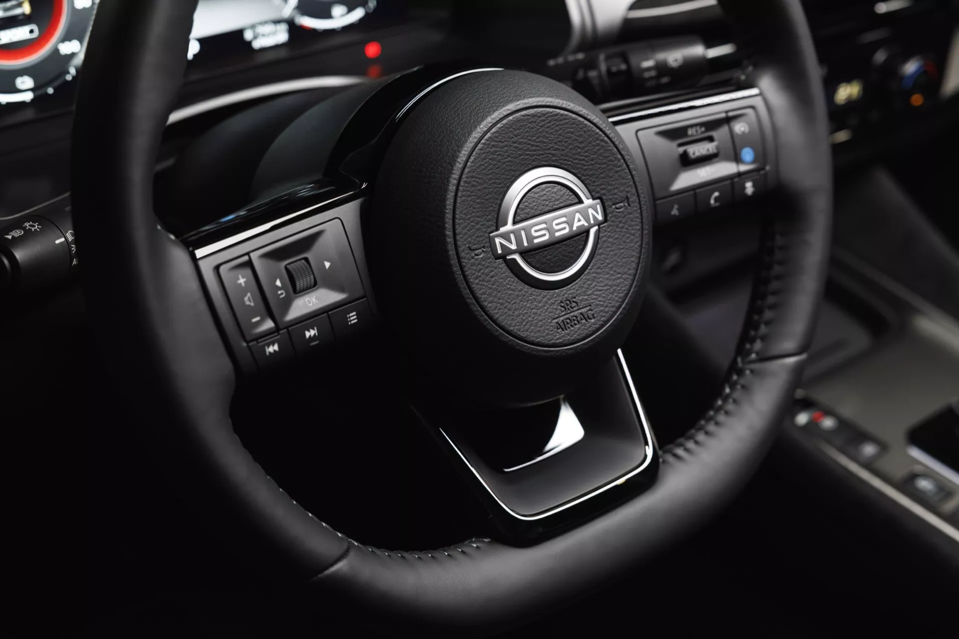 Interiér vozu Nissan Qashqai 2024 J12-facelift z pohledu řidiče s důrazem na moderní tříramenný volant potažený kůží s multimediálními a tempomatovými tlačítky. Uprostřed volantu je zobrazeno logo Nissan se sofistikovanými stříbrnými akcenty. Na obrázku je také částečně viditelný přístrojový panel, dotykový infotainment systém s lesklými černými lištami a tlačítky pro ovládání klimatizace, což ilustruje design kabiny zaměřený na řidiče.