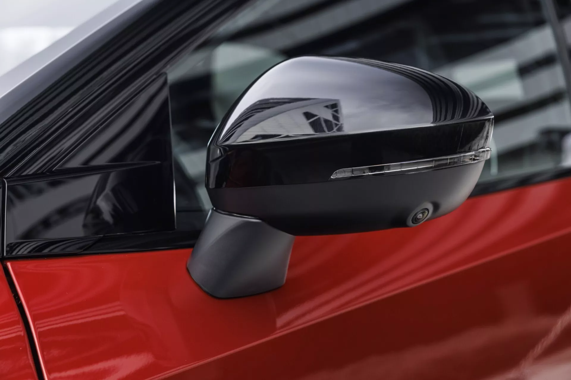 Detailní záběr bočního zrcátka moderního auta z bočního úhlu, zaměřený především na aerodynamický design zrcátka a integrovaný směrový ukazatel. Lesklé, reflexní černé pouzdro doplňuje živou červenou barvu karoserie, což odráží pečlivou rovnováhu estetické přitažlivosti a moderní automobilové technologie, konkrétně Nissan Qashqai 2024 (J12-facelift).