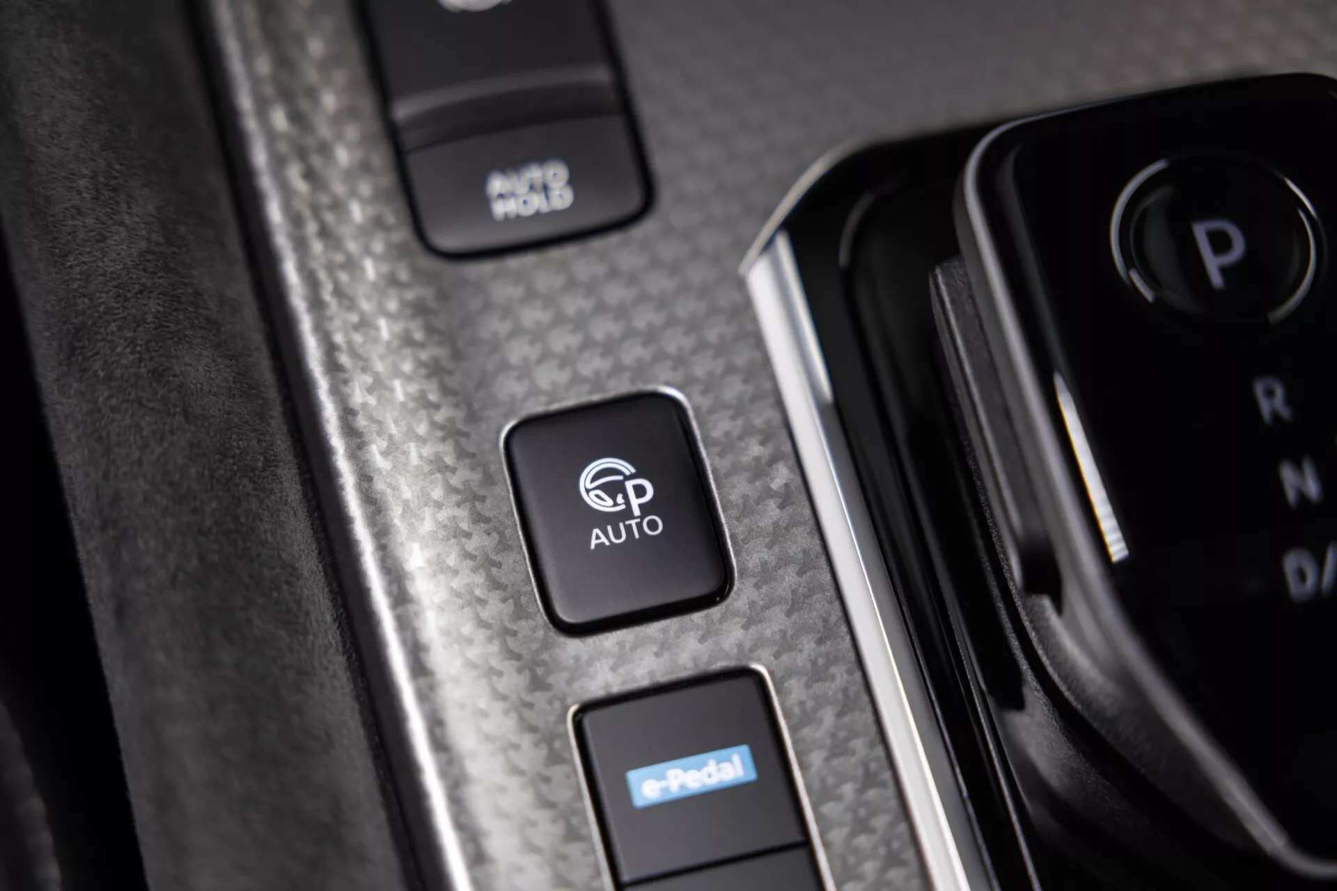 Detailní záběr ovládacích tlačítek na bočním panelu interiéru Nissan Qashqai 2024 (J12-facelift). Zaměřeno na tlačítko 'AUTO' s možnou funkcí automatického systému, tlačítko 'e-Pedal' pro elektronický pedálový systém jako jízda jedním pedálem a tlačítko 'ABS' pro systém proti zablokování kol. Texturovaný panel se vzhledem uhlíkových vláken nebo kartáčovaného kovu a lesklým lemováním kolem tlačítka 'AUTO'. Zdůrazňuje pokročilou techniku vozidla a asistenční systémy při řízení.