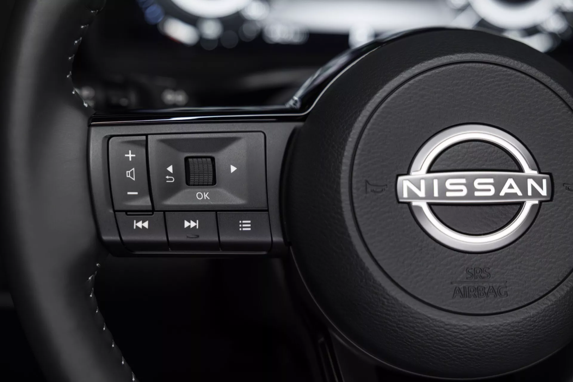 Detailní záběr na volant vozu Nissan Qashqai 2024 (J12-facelift) s ovládacími tlačítky na levé straně pro multimédia, včetně ovladačů hlasitosti, tlačítek pro navigaci mezi skladbami a tlačítkem pro potvrzení OK; logo Nissan uprostřed, rozmazané moderní interiérové pozadí s odrazy digitálního displeje, pohled z pozice řidiče nebo předního spolujezdce.