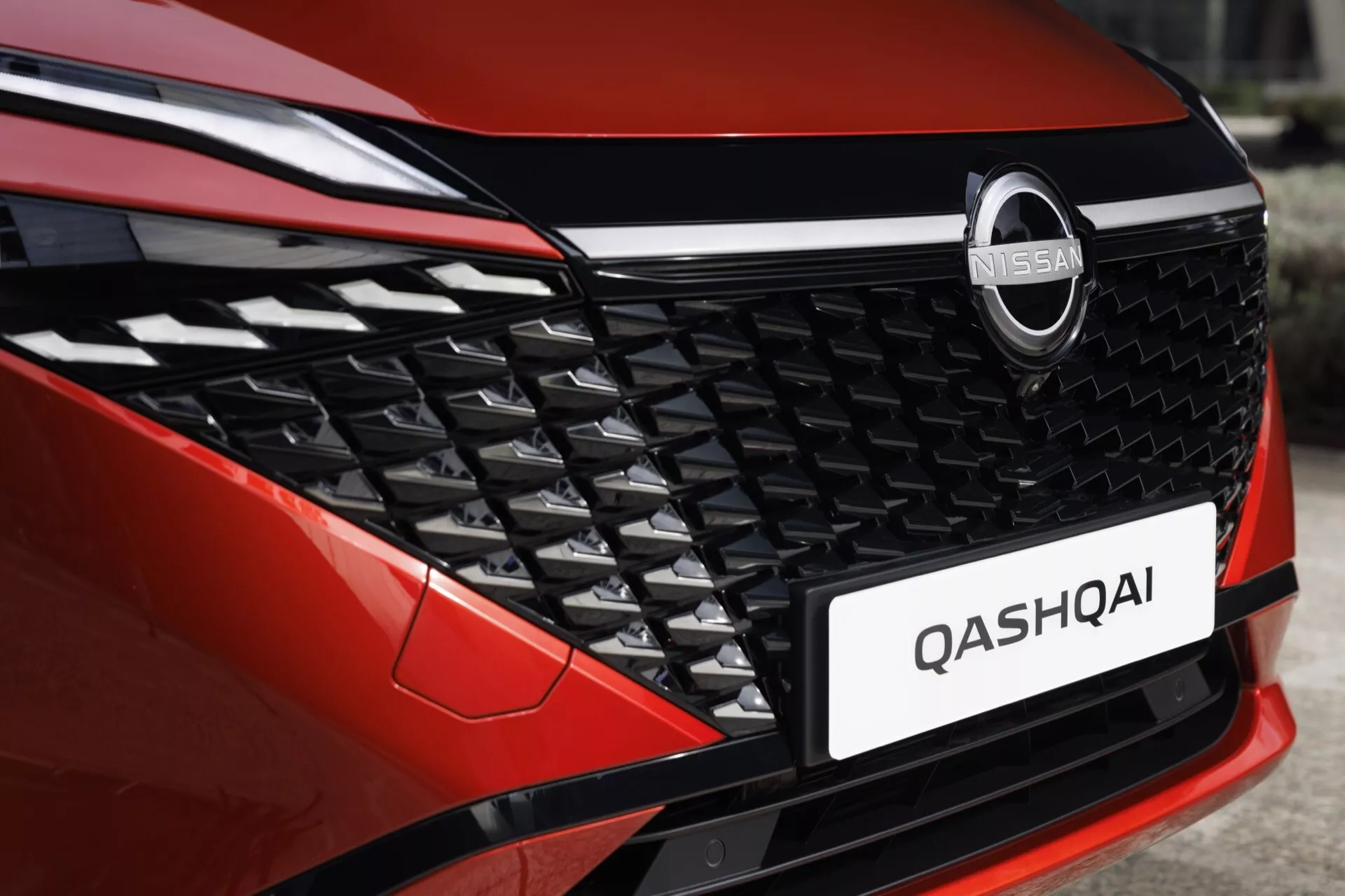 Detailní pohled na přední mřížku vozu Nissan Qashqai 2024 (J12-Facelift). Obrázek je pořízen ze šikmého úhlu a zvýrazňuje levou přední stranu s detailním pohledem na moderní, složitý design mřížky a část kapoty. Chromovaný znak Nissan je výrazně umístěn uprostřed, pod ním se nachází označení modelu 'QASHQAI'. Karoserie vozu má metalickou červenou barvu a spodní okraj ukazuje sportovní černý akcent na nárazníku. Přirozené osvětlení odhaluje jemné detaily designu a prvků vozidla.