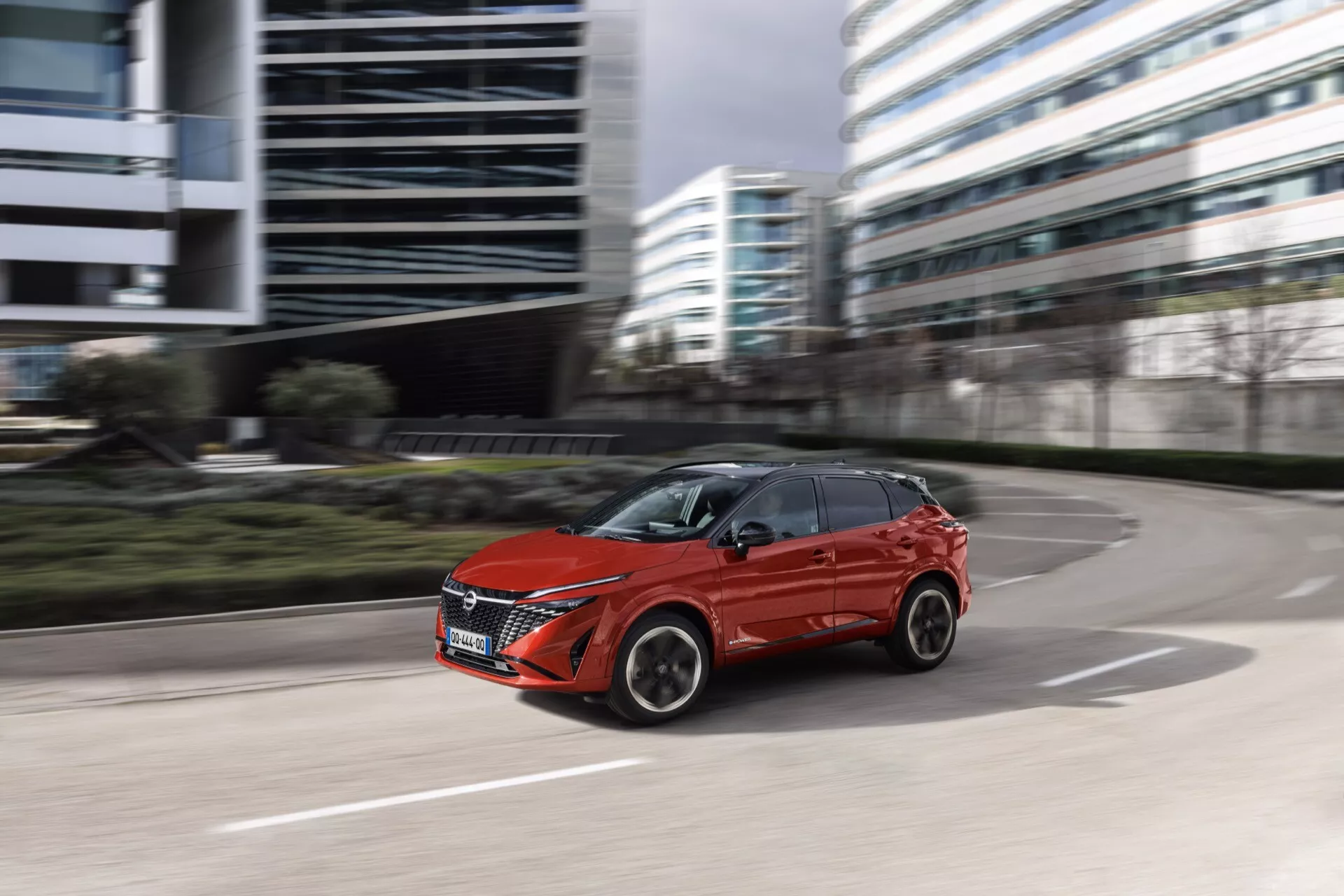 Dynamický obraz červeného Nissan Qashqai 2024 (J12-Facelift) v pohybu, s mírně rozmazaným pozadím naznačujícím rychlost. Zachycen z nízkého úhlu, vůz udržuje silnou přítomnost díky své diagonální poloze a rotujícím kolům. Zobrazen z bočně-předního perspektivu, výrazné rysy zahrnují elegantní LED nebo HID světlomety, složitě navrženou přední masku, vícepaprsková litá kola a aerodynamické tvarované linie karoserie. Městské pozadí s reflexními kancelářskými budovami naznačuje městský životní styl.