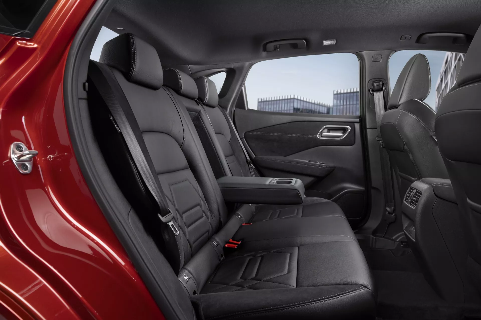 Interiér vozu Nissan Qashqai 2024 (J12-Facelift) zobrazující zadní prostor pro cestující pohledem z zadních dveří na straně řidiče. Černé kožené sedačky s prošitím, tvarované pro pohodlí a dostatek místa na nohy, vyjadřují sofistikovanost. Vybavení zahrnuje lavičku stylu sezení, nastavitelnou opěrku hlavy, chromovou klikou dveří, mřížku reproduktorů, ovládání elektrických oken, úložnou přihrádku, středovou konzoli s ventilačními otvory, jemné ambientní osvětlení, kobercové podlahy a integrované vývody pásů, což zdůrazňuje luxusní a dobře vybavené auto.
