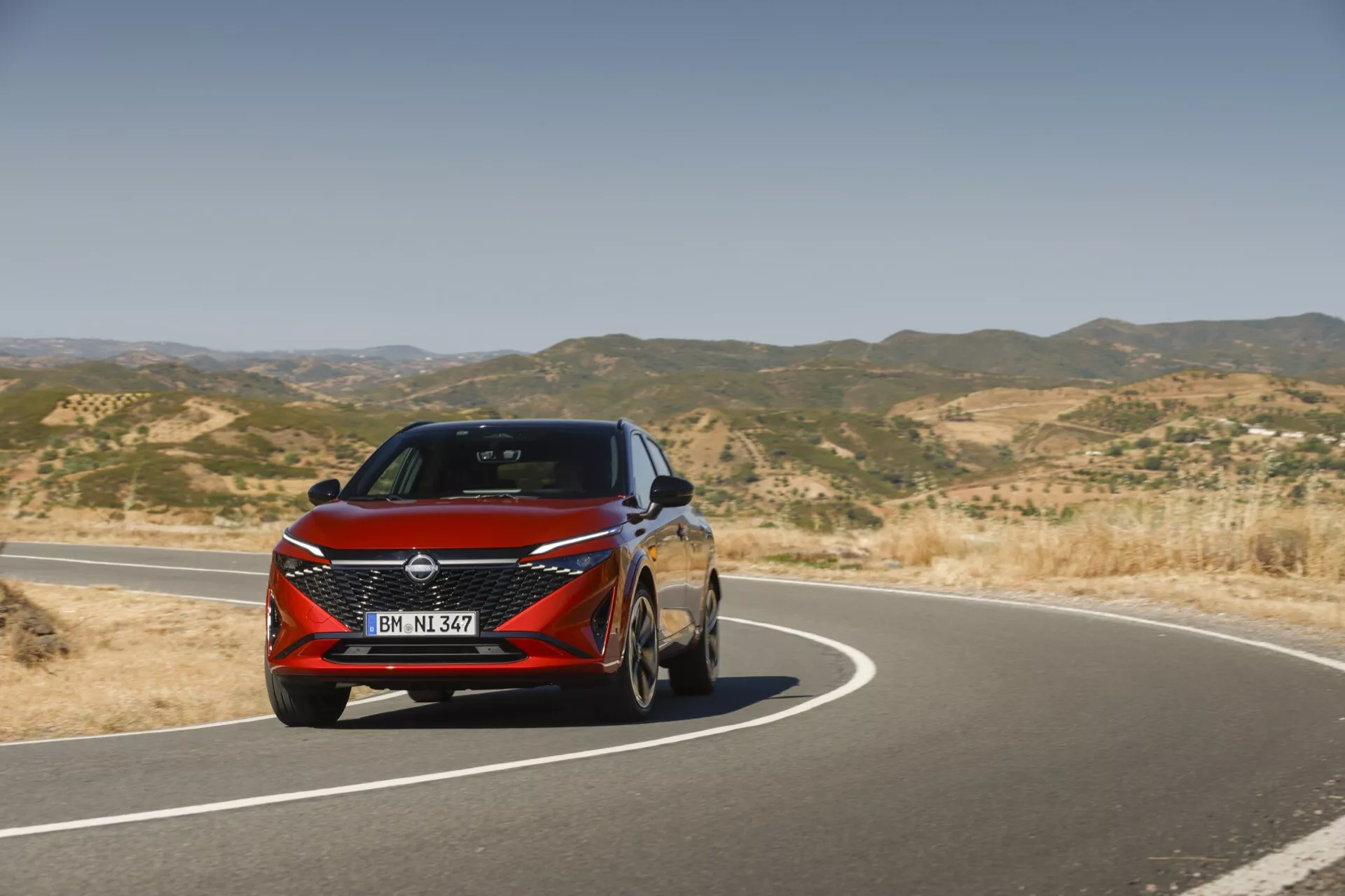Přední tříčtvrteční pohled na Nissan Qashqai 2024 (J12-facelift) s výraznou mřížkou chladiče, elegantními LED světlomety s integrovanými denními světly, tvarovanou kapotou, hliníkovými koly, aerodynamickými bočními zrcátky a metalickou lesklou povrchovou úpravou. Dynamické linie a křivky karoserie vyzařují moderní, sportovní vzhled. Auto je na fotografii v pohybu na zakřivené asfaltové silnici v suché, kopcovité krajině za jasného počasí.
