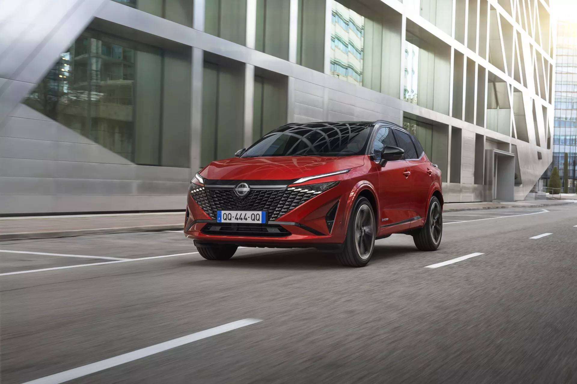 Moderní červený hatchback Nissan Qashqai 2024 (J12-facelift) zachycený v pohybu z trojčtvrtinového předního pohledu, se světlomety LED, výraznou mřížkou, tvarovanou kapotou, zrcátky s integrovanými LED směrovkami, pohybujícími se litými koly; městské prostředí s moderními budovami, denní osvětlení s vrženými stíny naznačujícími přirozené světlo shora.