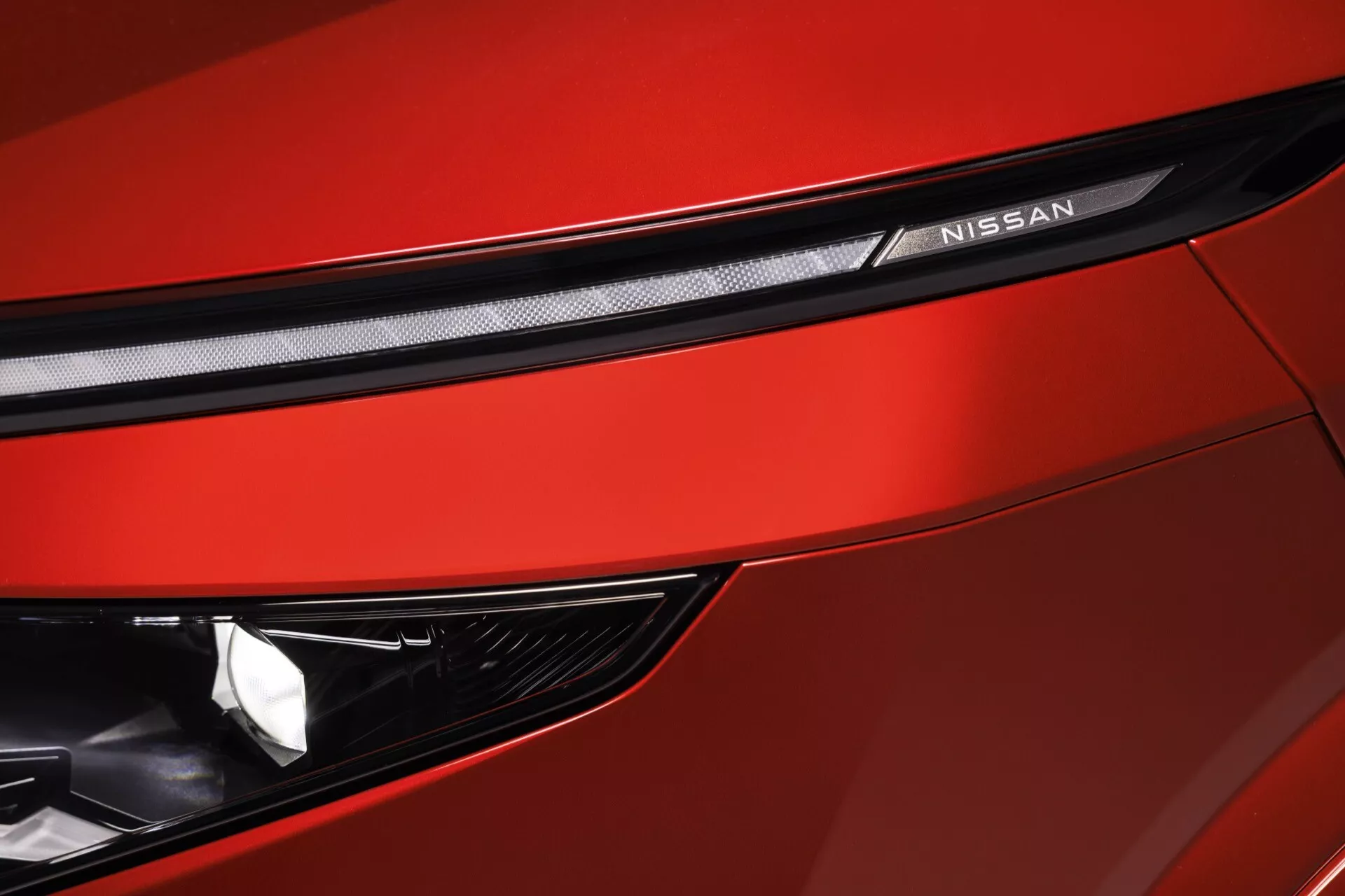 Detailní záběr na přední část červeného modelu Nissan Qashqai 2024 (facelift J12), zdůrazňující designové prvky a technologické vybavení. Záběr, pořízený pod mírným úhlem, zvýrazňuje část mřížky s elegantním, texturovaným vzorem, nápis 'NISSAN' na černém proužku nad mřížkou, svalnaté kontury kapoty, část elegantního úhlového světlometu pravděpodobně s LED nebo HID technologií a jasně červený lak naznačující sportovní výbavu.