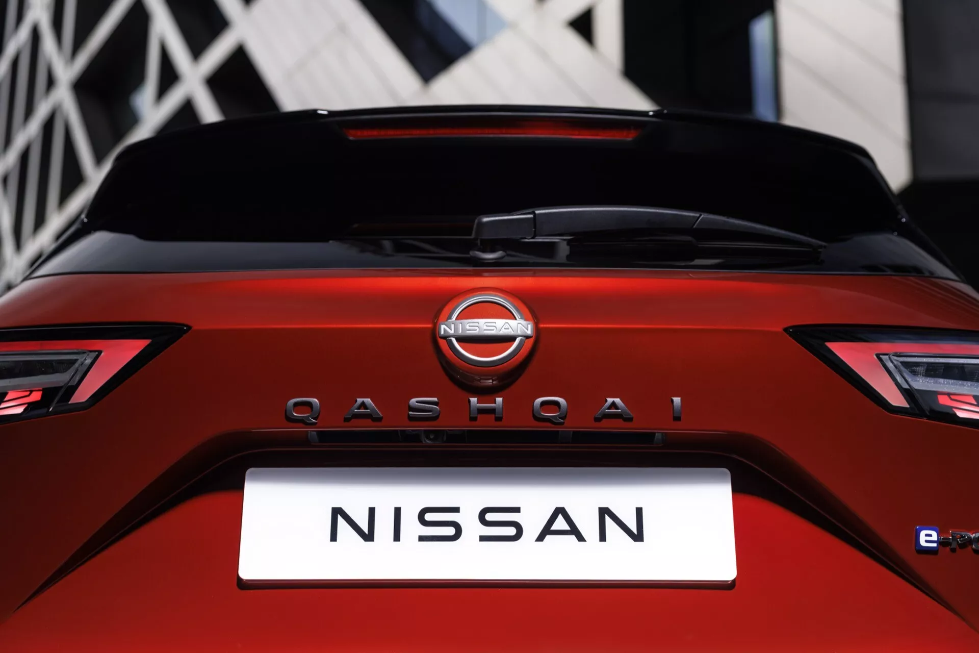 Zadní pohled na Nissan Qashqai 2024 J12-facelift, zdůrazňující oblast pátých dveří se zaměřením na zadní okno a boomerangová LED zadní světla. Záběr na výrazný znak Nissan nad názvem modelu QASHQAI s označením e-POWER naznačujícím hybridní systém. Vystaven v tmavě červené barvě s lesklým povrchem a ostrými designovými prvky. Městské pozadí, rozostřené.