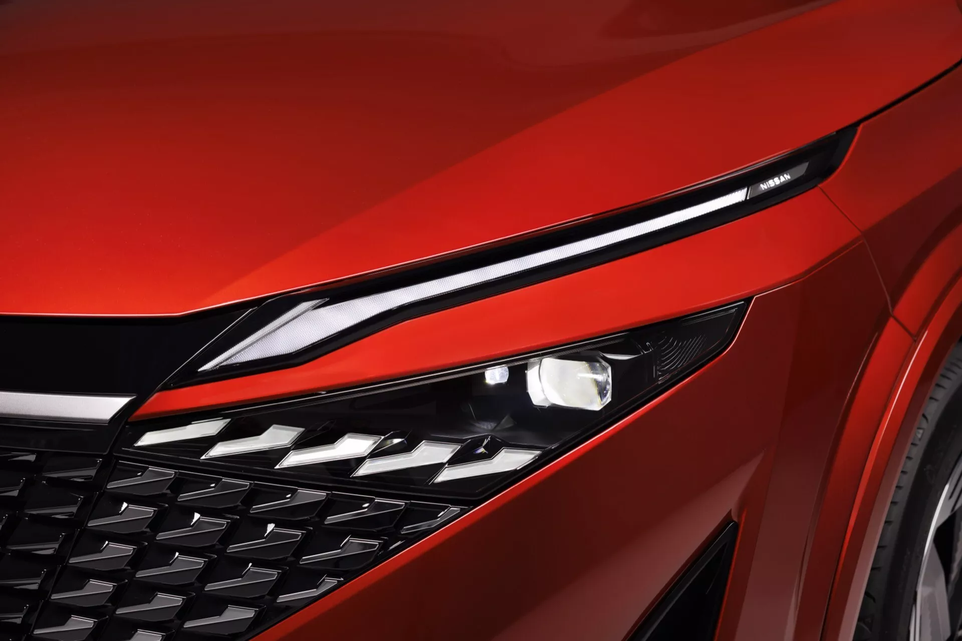 Detailní záběr na světlomet a mřížku červeného Nissan Qashqai 2024 (J12-facelift), zachycený z nízkého úhlu pro zvýraznění elegantního profilu a pokročilé LED osvětlovací technologie. Světlomet, úzká LED jednotka pravděpodobně s denními světly, je rozsvícený a demonstruje automatické osvětlovací funkce. Mřížka má diamantový nebo polygonální vzor, naznačující aktivní aerodynamiku nebo pokročilé chladicí systémy. Kovově červený panel karoserie se vyznačuje lesklým povrchem a sportovními konturami. Obrázek se soustředí na tyto prvky, s rozmazaným pozadím pro zdůraznění detailů a designu předního čtvrtpanelu.