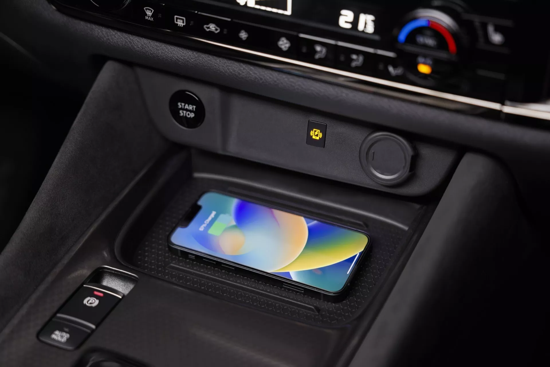 Interiér vozu Nissan Qashqai 2024 (J12-facelift) zobrazující přední pohled na středovou konzolu s bezdrátovou nabíjecí podložkou s chytrým telefonem, pokročilým klimatozací systémem s ukazateli '21' a 'AUTO', startovacím tlačítkem se žlutým varováním, elektronickou parkovací brzdou a částečně viditelnou pákou řadicí páky v interiéru z tmavého plastu s matným povrchem.