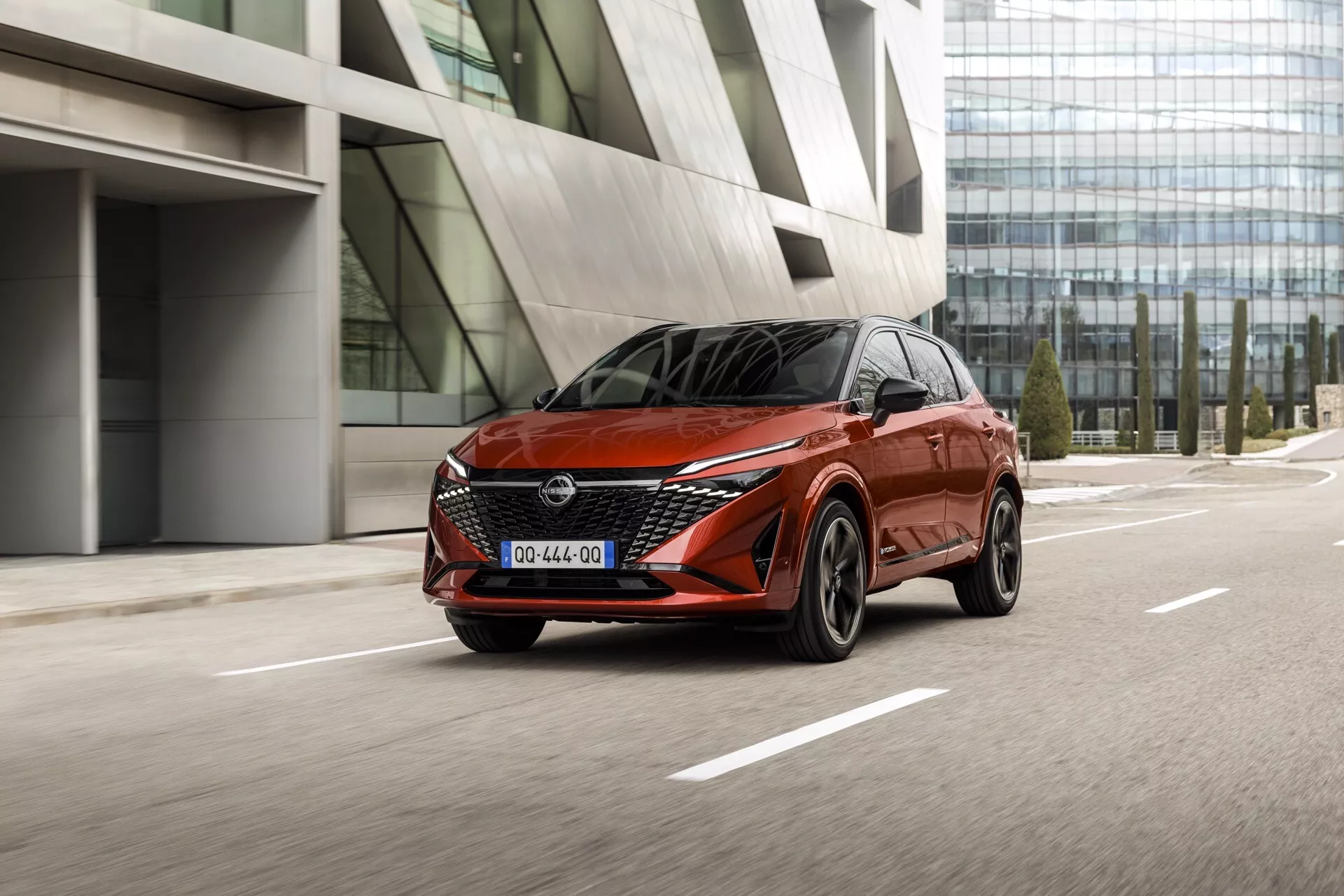 Nissan Qashqai 2024 (facelift J12) v pohybu, zachycen z nízkého úhlu ve tříčtvrtečním předním pohledu, zdůrazňující jeho sportovní a moderní designové linie s výraznou červenou barvou. Umístěný na levé straně rámu na pozadí městské architektury s reflexními skleněnými budovami. Zvýrazněny jsou elegantní LED světlomety auta, výrazná přední mřížka s logem Nissan, kola z lehkých slitin s vícepaprskovým designem a ostrá aerodynamická karoserie.