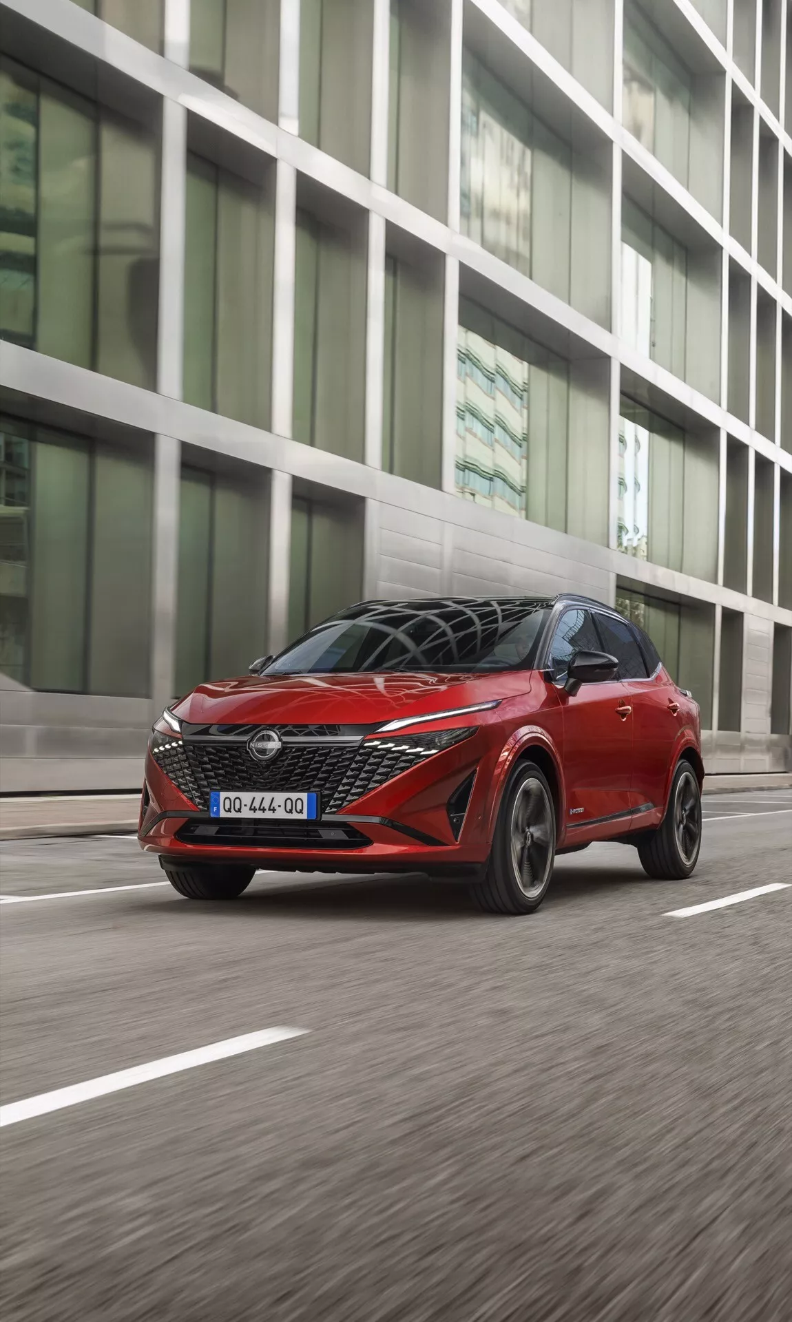 Dynamický snímek modelu Nissan Qashqai 2024 (J12-Facelift) v pohybu, pořízený z nízkého a lehce předního úhlu. Fotografie zachycuje přední levý bok vozu, zvýrazňuje charakteristický design mřížky s emblémem, elegantní LED světlomety, vícelistové slitiny kol, boční zrcátka s možnými integrovanými směrovkami a kovový lak. Městské pozadí je lehce rozmazané, což zdůrazňuje rychlost a pohyb vozu. Sportovní linie střechy a rozšířená kabina svědčí o crossoverovém tělesi vozu. Pravděpodobně se používá k reklamě, prezentuje design a výkon.