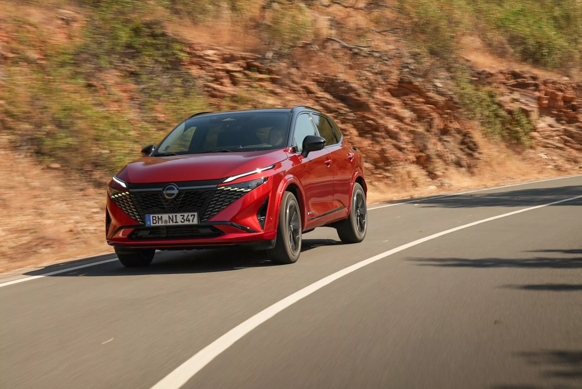 Obrázek zachycuje dynamický boční pohled na červený Nissan Qashqai 2024 (facelift J12) jedoucí po zakřivené silnici. Fotografie pořízená z mírně předního úhlu zvýrazňuje řidičovu stranu, ukazuje přední levou čtvrtinu s řidičovými dveřmi, částí kapoty, předním kolem, blatníkem a částí světlometu. SUV se vyznačuje ostrými a hranatými liniemi, které zdůrazňují jeho moderní a agresivní design. Viditelná je velká, charakteristická přední mřížka, úzké hranaté světlomety a pokročilá osvětlovací technologie včetně LED denních světel a mlhovek. Moderní vícepaprskový design kol naznačuje sportovní výbavu. Vozidlo je zobrazeno za denního světla s rozmazaným pozadím naznačujícím pohyb, což je výsledek techniky panning použité fotografem.