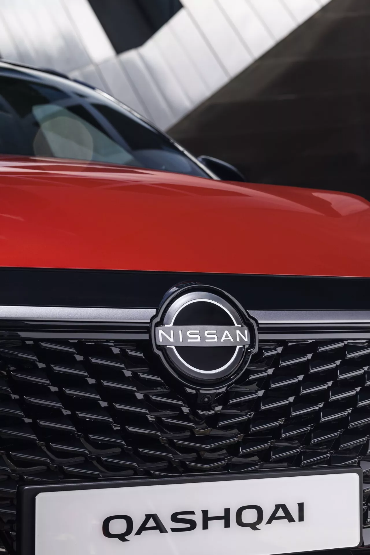 Zblízka přední část vozu Nissan Qashqai 2024 (J12-Facelift). Fotografie, pořízená z předního kvadrantového úhlu, zvýrazňuje výrobní emblém prominentně umístěný ve středu propracované moderní mřížky s propleteným 3D mřížkovým designem. Jméno 'QASHQAI' je jasně viditelné na oblasti registrační značky. Kapota ukazuje živou červenou dvoutónovou barevnou schéma s tenkou černou lištou, zdůrazňující prémiový design vozu. Malá hloubka ostrosti udržuje zaměření na tyto klíčové prvky a rozmazává pozadí.