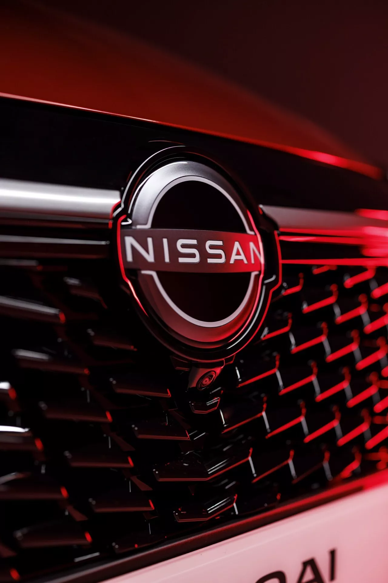 Detailní záběr na přední mřížku vozu Nissan Qashqai 2024 (J12-Facelift) s kovovým, chromovaným logem Nissan uprostřed, umístěným na mřížce s hexagonálním vzorem. Záběr je pořízen z mírně mimo střed, z pravého úhlu s malou hloubkou ostrosti, zdůrazňující logo a design mřížky. Světlo vytváří odrazy a jemný lesk po okrajích, zvýrazňující prémiovou kvalitu vozu v tmavých tónech s rozmazaným červeným pozadím.