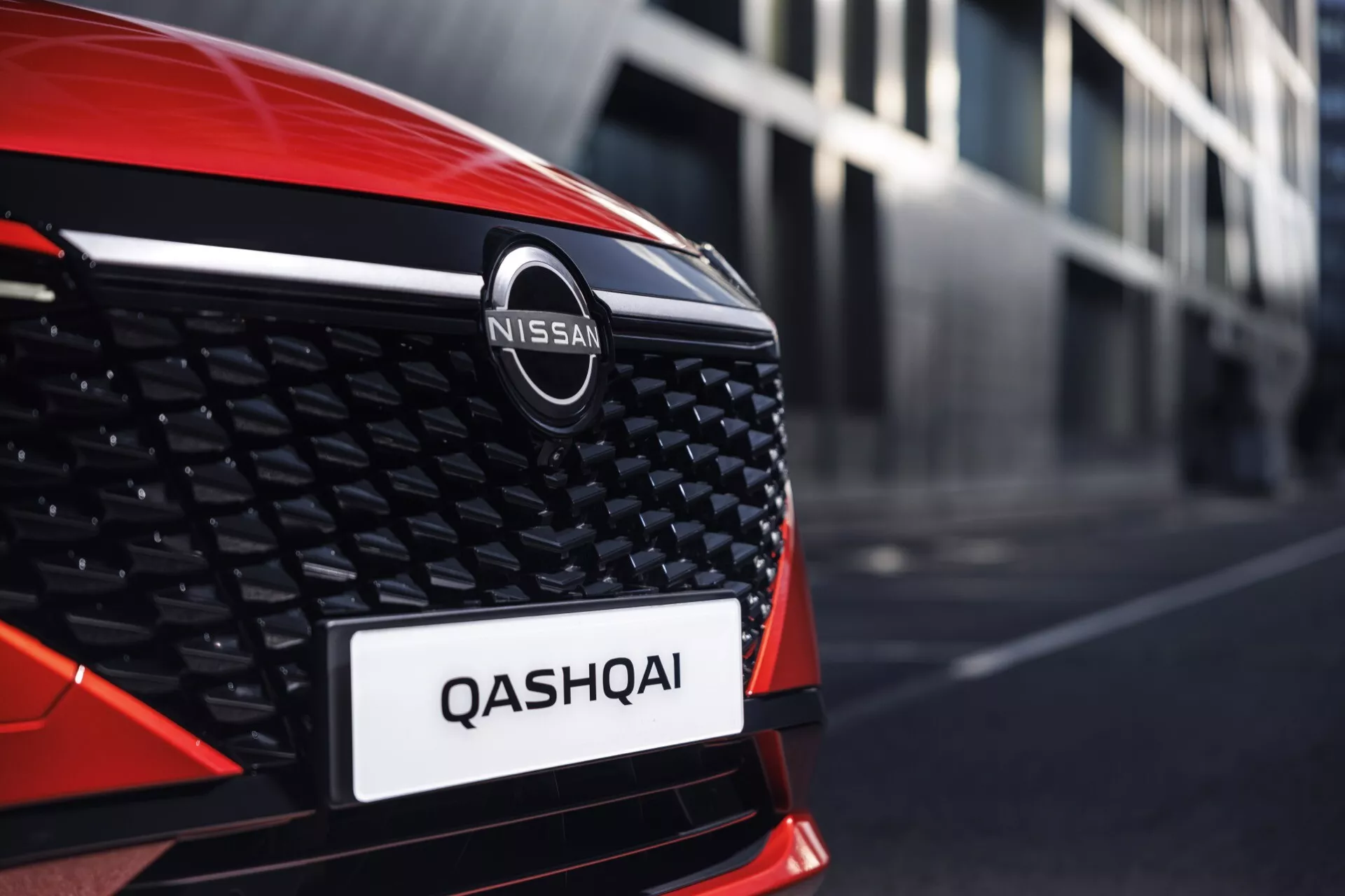 Detailní pohled z úhlu na přední část levého boku červeného Nissanu Qashqai 2024 (J12-facelift). Obrázek se zaměřuje na oblast mřížky s výrazným logem Nissan, velkou chromovanou V-tvarovanou lištou, voštinovým designem a viditelným nápisem 'QASHQAI' na registrační značce. Lesklá černá mřížka kontrastuje s kovovým odznakem a jasně červenou barvou.