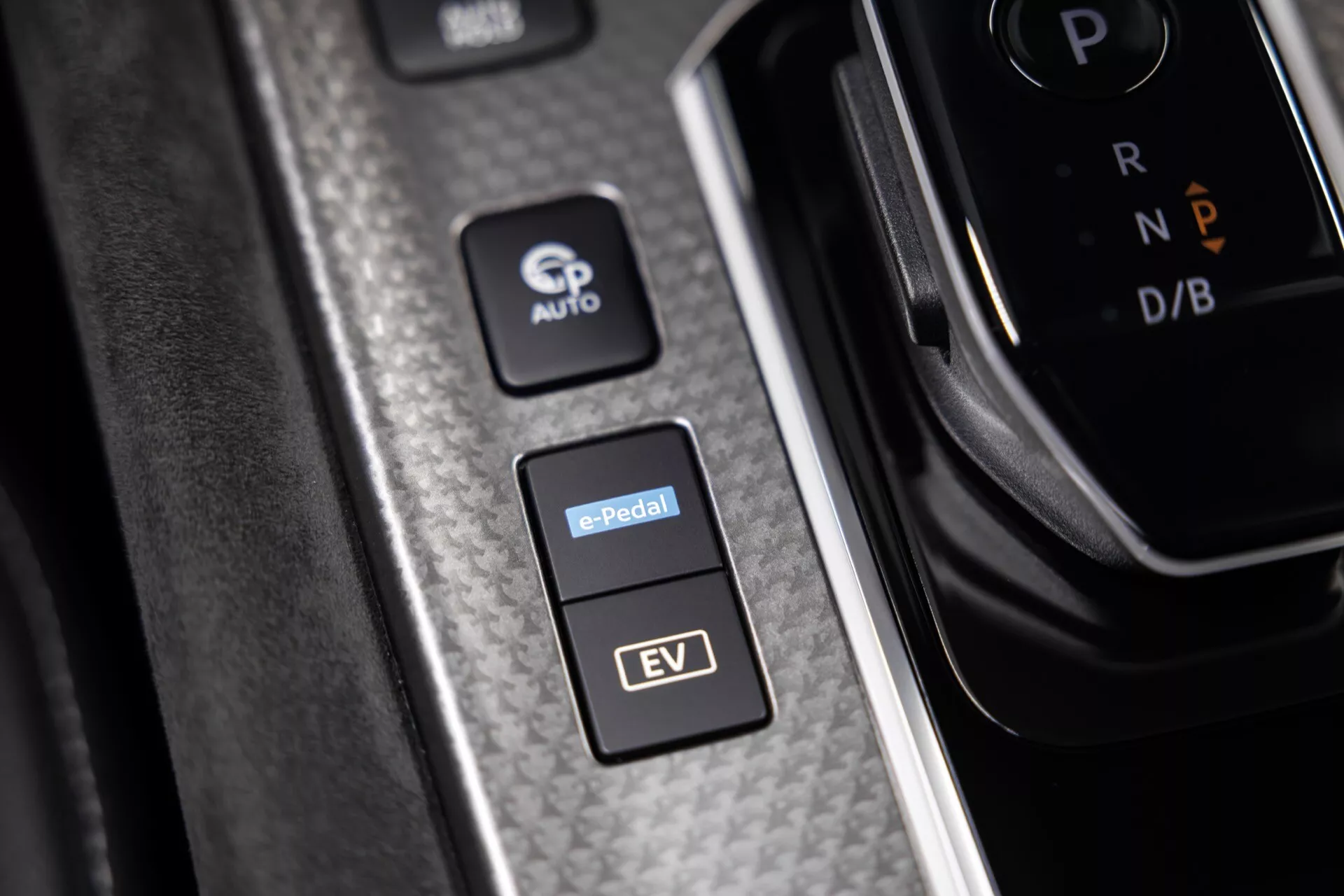 Detailní pohled na středovou konzoli vozu Nissan Qashqai 2024 (J12-facelift) z mírně zvýšeného úhlu, zvýrazňující karbonově-texturovaný povrch a tři odlišná tlačítka: 'P AUTO' pro automatické parkování nebo podržení, další s nápisem 'e-Pedal' pro jízdu s jedním pedálem v elektrických vozidlech a 'EV' označující elektrické schopnosti vozidla. Vpravo je částečně rozostřená řadicí páka s nápisem 'P' znamenající parkování, naznačující automatickou převodovku. Ideální pro diskusi o automobilové technologii, elektrických vozidlech a moderním designu interiéru vozu.