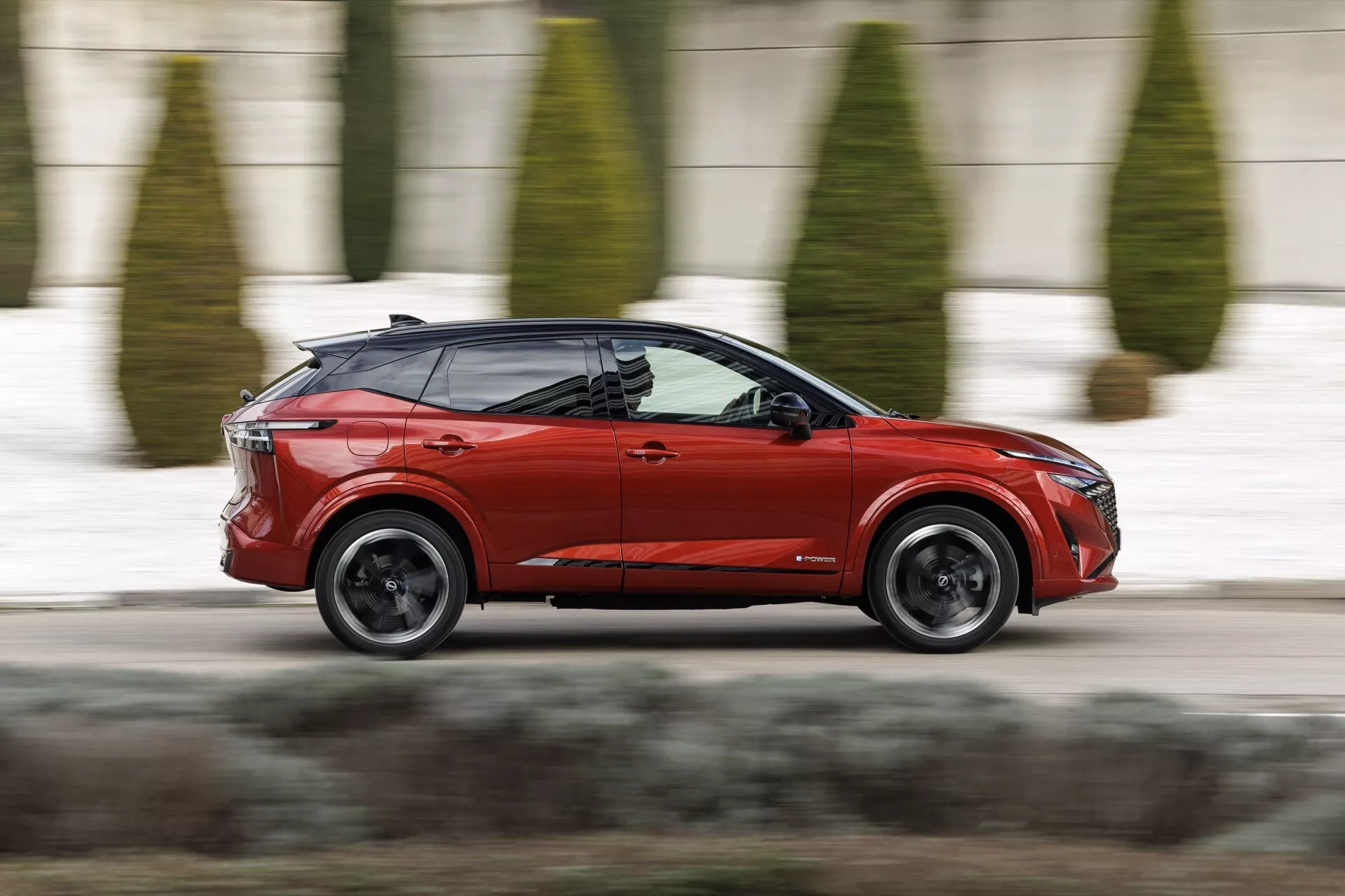 Dynamický boční profil moderního červeného crossoveru SUV Nissan Qashqai 2024 J12-Facelift v pohybu na městské ulici. Design vozidla zahrnuje ostrá LED přední světla, výraznou masku, bokozrcátka v barvě karoserie, svažující se střechu, výrazné blatníky s tmavými slitinovými koly, hladce integrované dveře, viditelnou panoramatickou střechu, černé sloupky vytvářející efekt plovoucí střechy, LED zadní světla s charakteristickým světelným podpisem a malý zadní spoiler. Technika sledování zachycuje auto v ostrém zaostření proti rozmazanému městskému pozadí, což naznačuje rychlost a dynamiku.