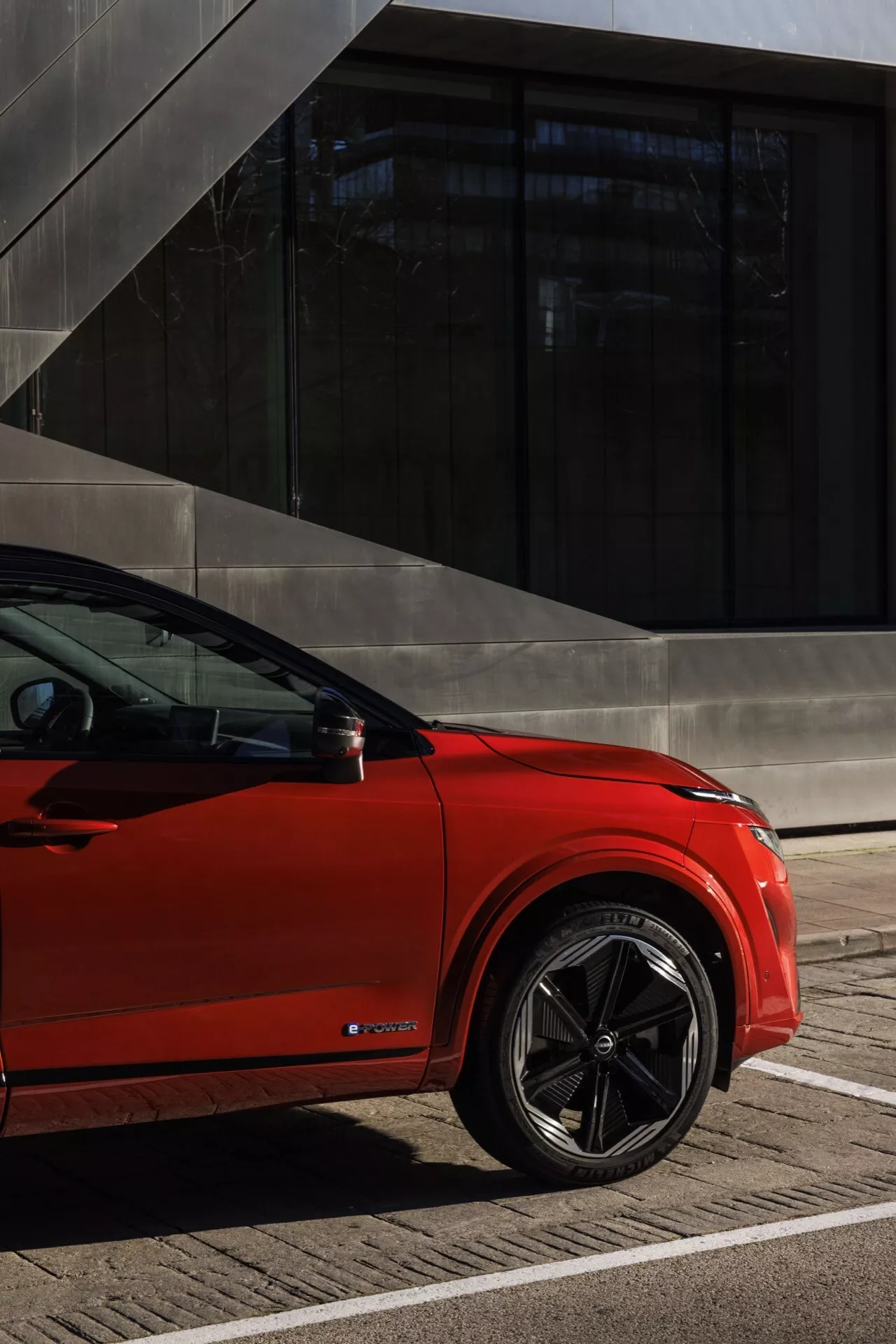 Nissan Qashqai 2024 (J12-facelift) v živé červené barvě, zachycen z nízkého úhlu, který zdůrazňuje sportovní zadní čtvrtinu a design vícepaprskových slitinových kol. Přirozené denní světlo zvýrazňuje hluboký zadní blatník, nízkoprofilové pneumatiky a elegantní linie zadních dveří, nárazníku a okna. Značka 'E-TRON' naznačuje elektrický model. Na pozadí moderní architektury s tmavými skleněnými okny a ostrými konstrukcemi tento obrázek podtrhuje prémiovou, současnou estetiku vozidla.