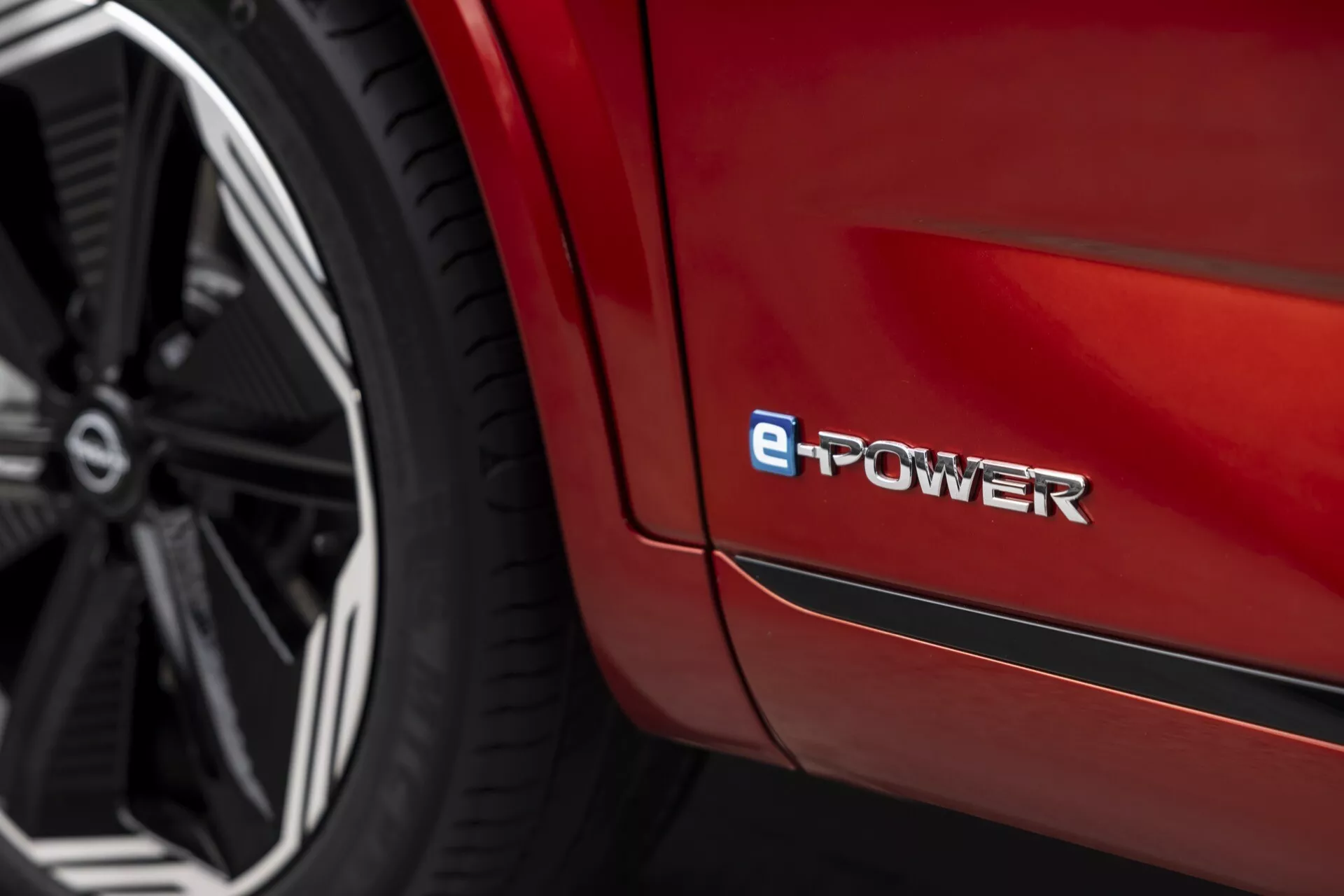 Detailní, šikmý pohled na část Nissan Qashqai 2024 (J12-facelift), zvýrazňující zadní blatník nad zadním kolem. Zářivý červený lesklý povrch s moderním, vícepaprskovým designem kola z lehkých slitin v tmavém provedení. Odznak 'e-POWER' označující elektrifikovaný pohonný systém. Zaměření na elegantní linie a křivky karoserie, s mělkou hloubkou ostrosti zvýrazňující odznak a rozmazané okolí.