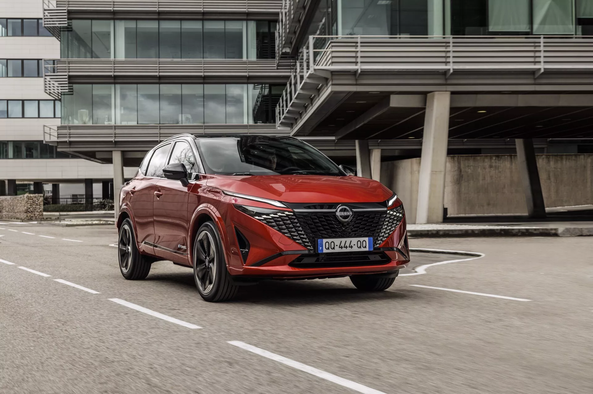 Moderní vůz Nissan Qashqai 2024 (J12-facelift) z pohledu zepředu z tříčtvrtečního úhlu, zvýrazňující přední mřížku s geometrickým vzorem, ostré LED světlomety, vícepaprsková hliníková kola, aerodynamická boční zrcátka s integrovanými indikátory směru a senzory u předního kola naznačující pokročilé asistenční systémy řidiče (ADAS). Automobil s agresivním a sportovním designem je zaparkován na vyznačeném zpevněném místě s moderní architekturou v pozadí, což naznačuje spojení s městským, současným životním stylem.
