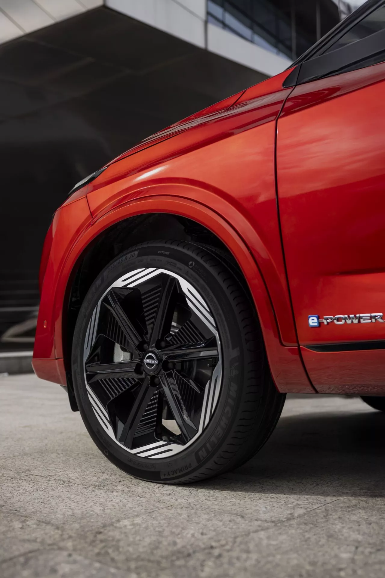Detailní zadní boční pohled na červený Nissan Qashqai 2024 J12-Facelift, zaměřený na složitý design kola s více paprsky a označení 'e-POWER', naznačující elektrifikovaný pohon. Nízký úhel záběru zdůrazňuje design kola s černými a strojově čelními paprsky, nízkoprofilovou pneumatiku a živou červenou kvalitu laku, zachycující moderní aerodynamický estetický vzhled.
