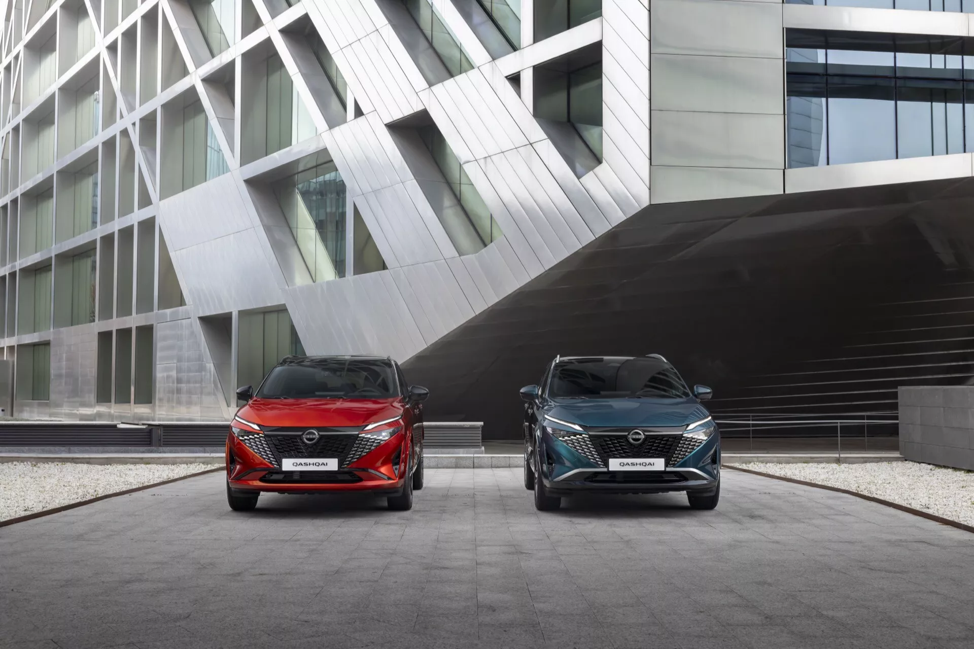 Nízký úhel záběru dvou modelů Nissan Qashqai 2024 (J12 Facelift) zaparkovaných před moderní budovou se skleněnou a hranatou architekturou. Oba vozy mají elegantní světlomety, výrazné mřížky chladiče a tvarované přední nárazníky s přívody vzduchu. Vozidla jsou umístěna pod mírným úhlem, což ukazuje na jejich symetrický design a technologické detaily přední části, jako jsou LED světlomety a přední parkovací senzory.