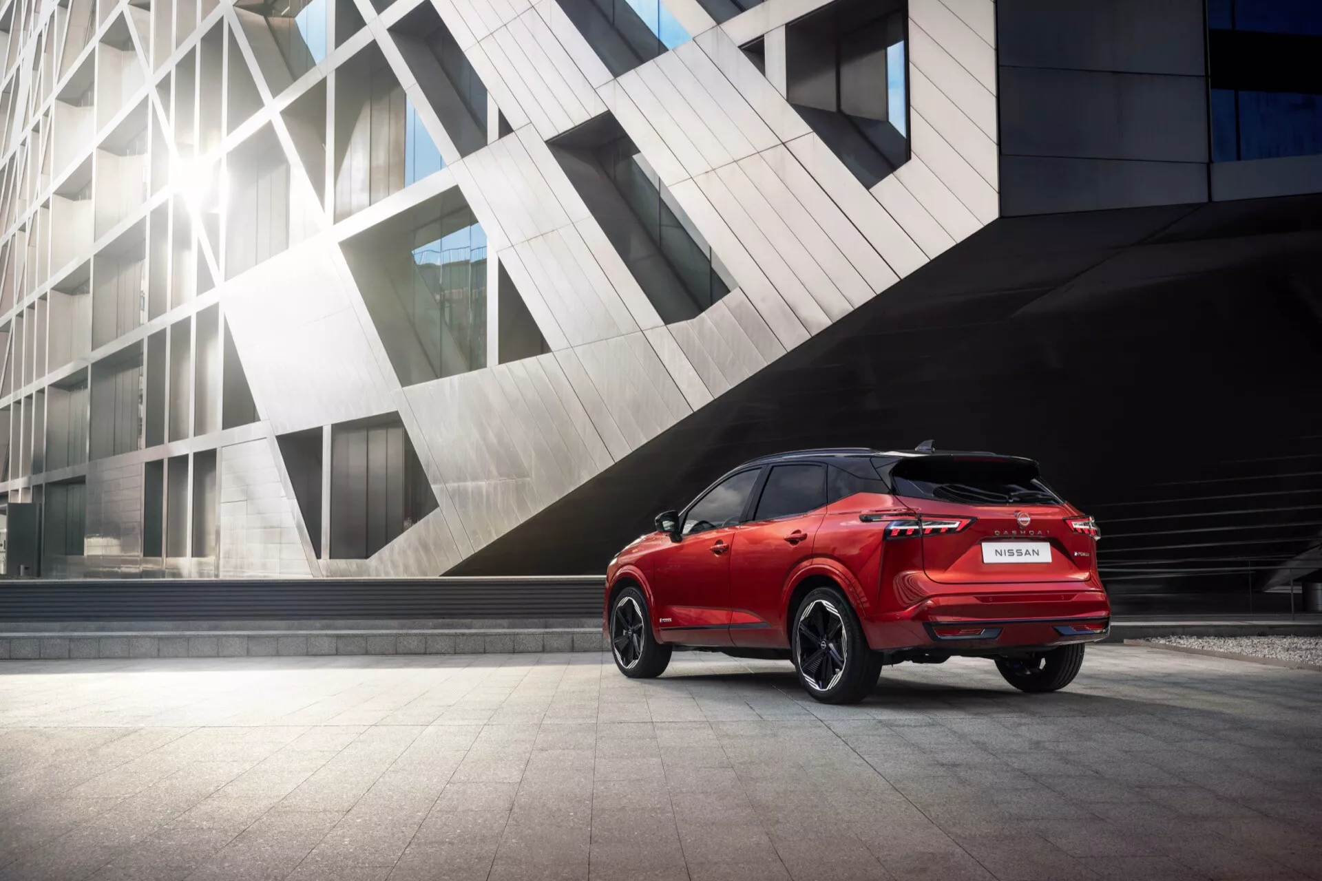Tříčtvrteční zadní pohled na červený Nissan Qashqai 2024 (J12 facelift), zaparkovaný na hladkém chodníku vedle moderní architektonické stavby. Obrázek zdůrazňuje LED zadní světla, zadní spoiler, žraločí anténu, velká vícepaprsková hliníková kola, branding a zadní modelové označení, zadní kameru a parkovací senzory, bočně zbarvená zrcátka, okna s tmavým tónováním a elegantní designové linie, vše zachyceno z mírně nízkého úhlu pro dynamický propagační vzhled.
