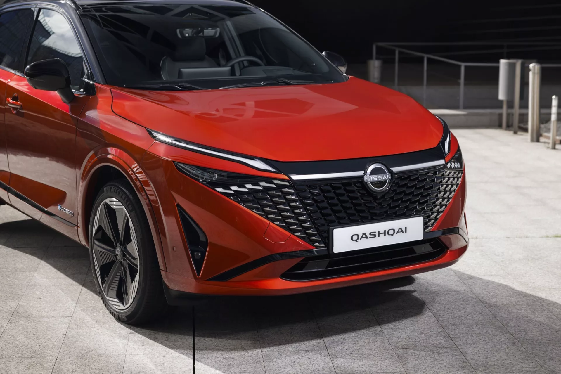 Obrázek ukazuje Nissan Qashqai 2024 (facelift J12) z nízkého předního tříčtvrtečního úhlu, zvýrazňující levý přední bok vozu. Auto, zaparkované v městském prostředí, má nápadný červený lak, který zdůrazňuje jeho kontury. Výrazný chromový akcent ve tvaru písmene V na velké přední mřížce, složitý vzor mřížky, elegantní LED světlomety a integrovaná denní světla zvyšují jeho moderní design. Sochovaná kapota a částečně viditelné zpětné zrcátko s integrovanými směrovkami přidávají sportovní vzhled. Kola naznačují design z lehkých slitin s více paprsky, což doplňuje moderní vzhled vozidla.