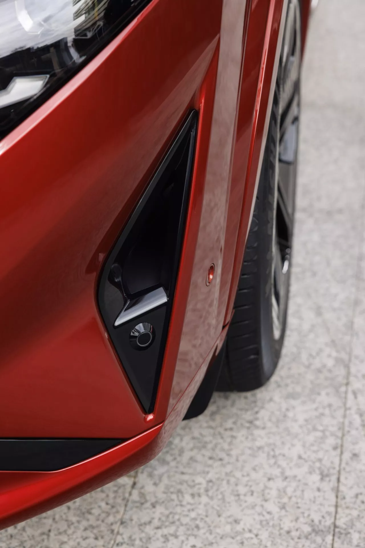 Detailní fotografie Nissan Qashqai 2024 (J12 facelift) zaměřená na přední levou stranu, zachycuje část elegantního, pravděpodobně LED světlometu, hladkou linii blatníku a hranatý černý nasávací otvor nebo větrací otvor na nárazníku s umístěním pro parkovací senzor. Obrázek ukazuje část moderně navrženého kola a zobrazuje černočervený nebo kaštanový lak automobilu, zdůrazňující těsné a rovnoměrné mezery mezi panely, což naznačuje kvalitní montáž a inženýrství. Dynamický diagonální úhel prezentuje hloubku a náhled na současný design estetiku vozidla.