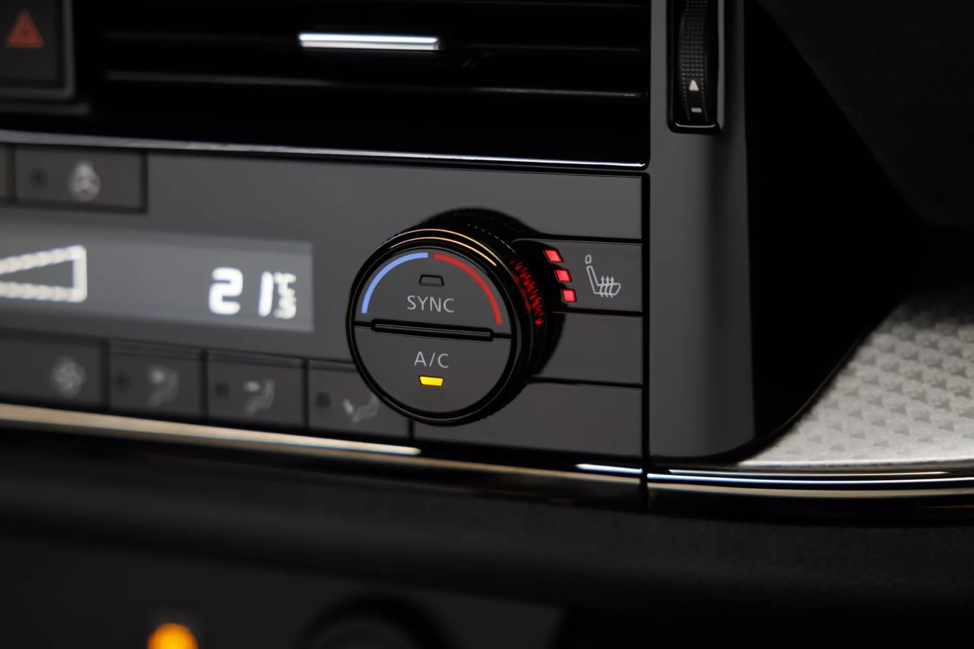 Detailní pohled na interiér modelu Nissan Qashqai 2024 (J12 facelift), který zvýrazňuje systém klimatizace umístěný na straně spolujezdce středové konzoly. Ústředním prvkem je ovladač klimatu nastavený na 21 stupňů, obklopený tlačítky jako 'SYNC' pro dvouzónovou klimatizaci a tlačítkem A/C, obě zvýrazněná červeným kontrolním světlem. Palubní deska má černý povrch s karbonovým vzorem a chromovou akcentní linkou, zatímco nad ovladačem je digitální displej s částečně viditelnými čísly '2 13'. Mělká hloubka ostrosti se zaměřuje na ovladač, čímž zdůrazňuje pokročilou a uživatelsky přívětivou klimatizační technologii vozu.