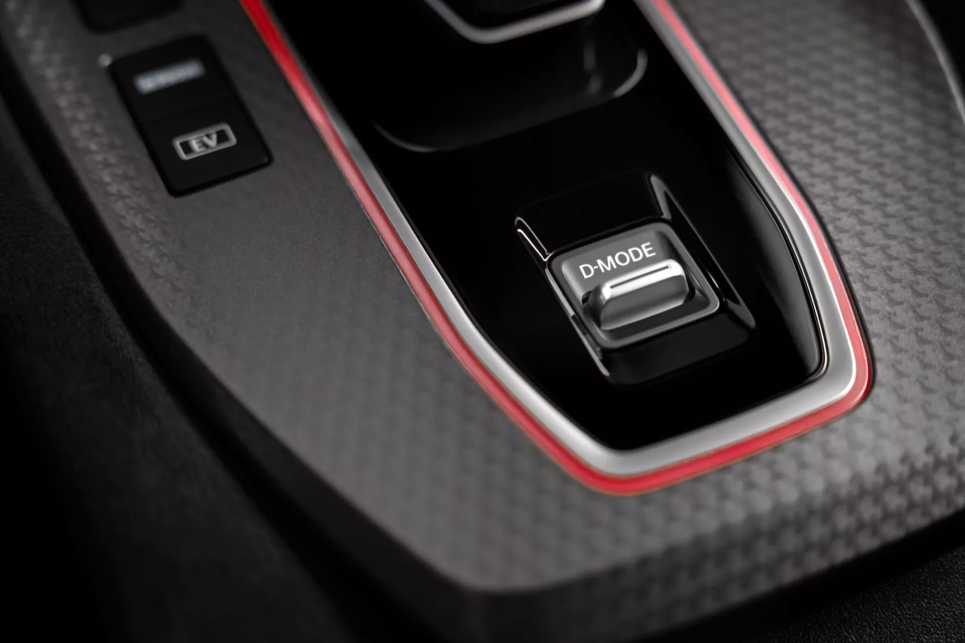 Detailní pohled na středovou konzoli interiéru Nissan Qashqai 2024 (J12-facelift), která zobrazuje volič převodovky označený 'D-MODE' umístěný v panelu s uhlíkovým vzorem a lesklým povrchem s červeným akcentem. Design naznačuje sportovní výkon. Konzola také obsahuje dvě tlačítka, jedno naznačující stabilitu nebo dynamiku vozidla a druhé pravděpodobně pro spuštění/vypnutí motoru.
