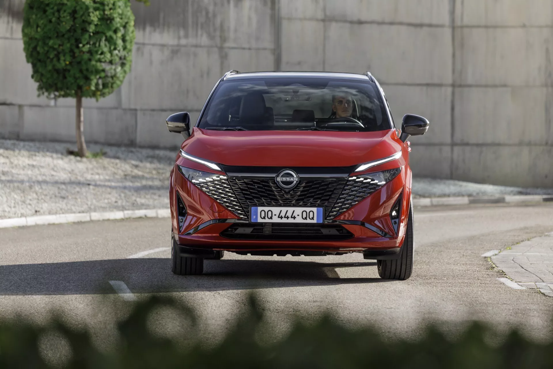 Nízký úhel 2024 Nissan Qashqai J12-facelift přijíždějící čelní pohled, předvádějící moderní design s výrazným V-tvarovým chromovaným akcentem na úhledné přední mřížce, ostrými LED světlomety a sportovním předkem. Zachycený v dynamickém městském prostředí, obraz zdůrazňuje aerodynamické kontury, senzory pro asistenci řidiče a široké čelní sklo, naznačující prostorný interiér.