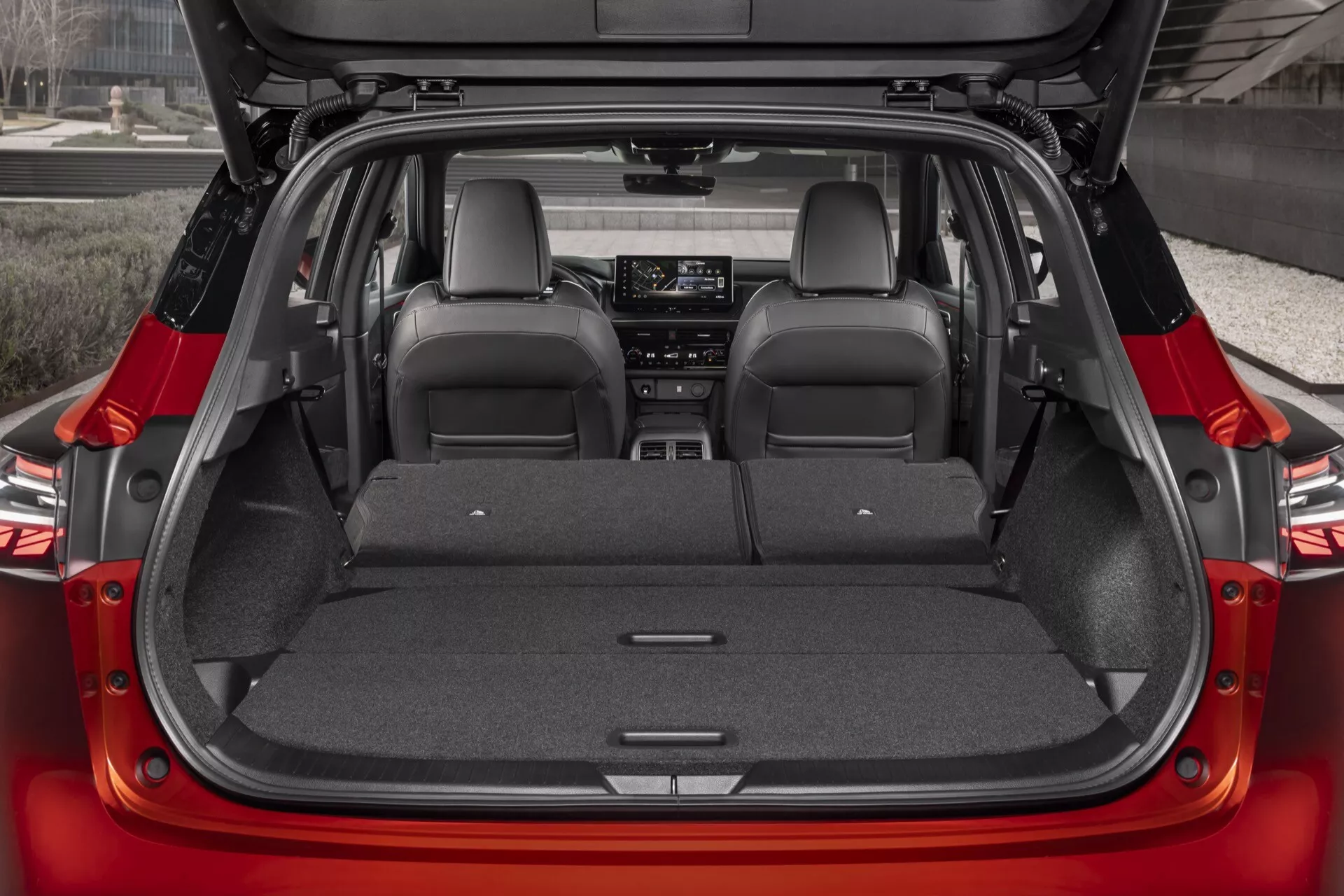Zadní interiér červeného vozu Nissan Qashqai 2024 (J12-facelift) zobrazený zvenku, ukazující prostorný otevřený kufr se zvednutým zadním víkem. Kamera nabízí střední, přímý záběr zdůrazňující sklápěcí zadní sedadla, poskytující dostatek úložného prostoru. Nákladní prostor má tmavě tónovanou šedou nebo černou kobercovou podložku. Viditelné designové prvky zahrnují výrazné kožené čalounění, které naznačuje luxusní úrovně výbavy, a částečně viditelný infotainmentový systém pravděpodobně vybavený moderními technologiemi. Otevřený kufr může naznačovat elektricky ovládané víko zavazadlového prostoru, zdůrazňující pohodlí.