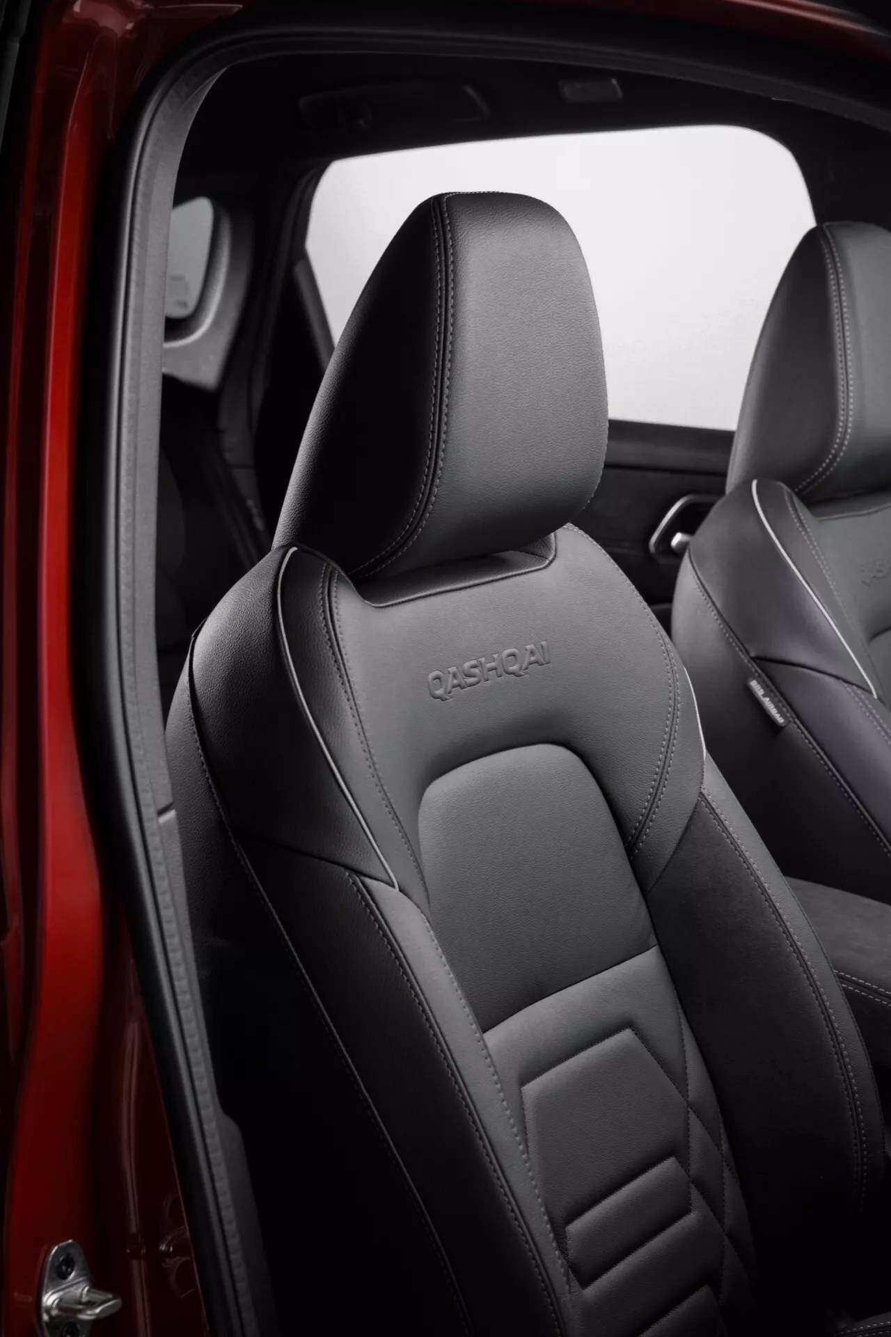 Detailní záběr interiéru předních sedadel vozu Nissan Qashqai 2024 J12-facelift zaměřený na sedadlo řidiče. Černé kožené čalounění s propracovaným šitím a embosované logo 'QASHQAI' na horní části sedadel vyzdvihují luxusní a sportovní design.