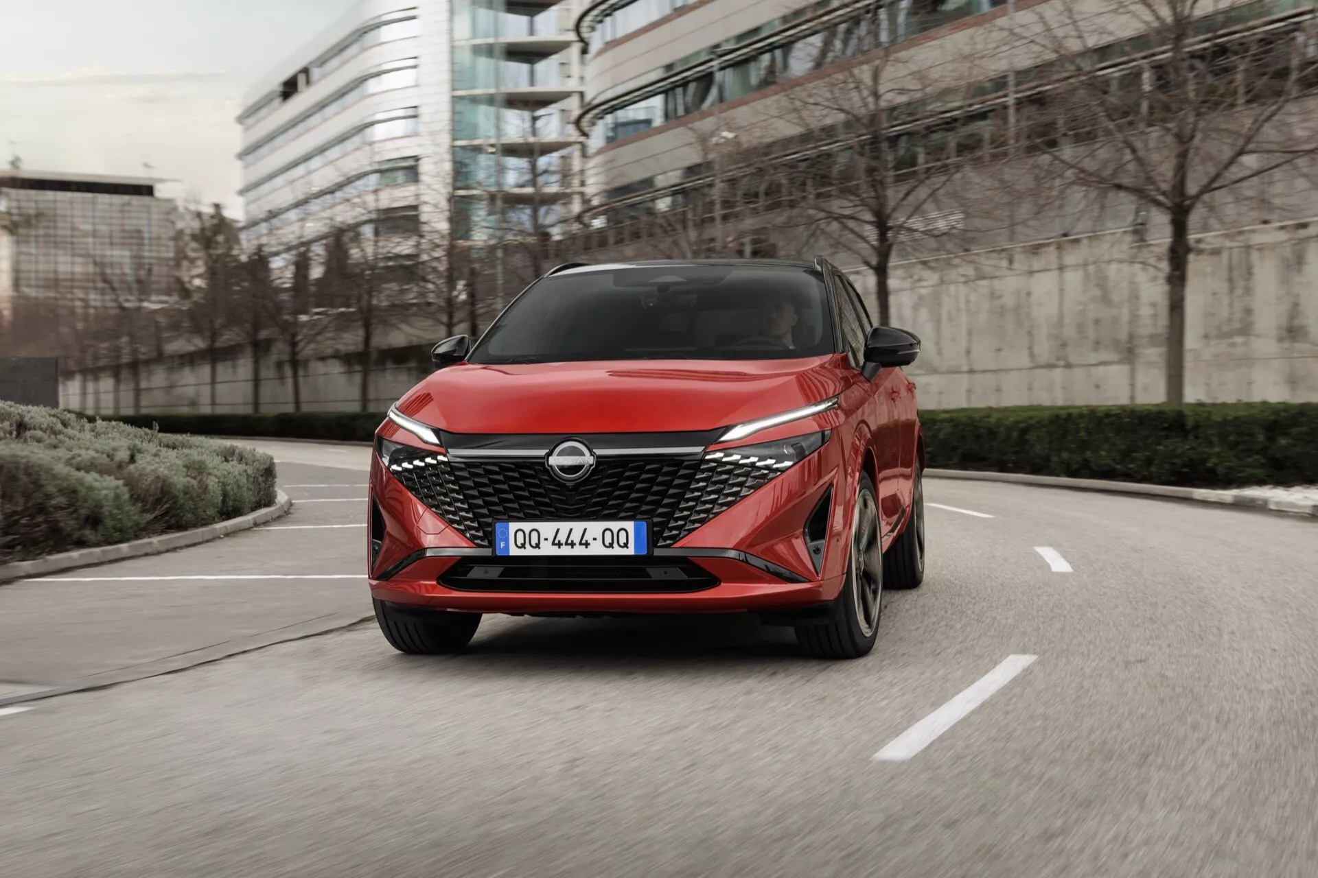 Dynamický pohled zepředu na Nissan Qashqai 2024 (J12-Facelift) v červené barvě, s nízkým úhlem zvýrazňujícím agresivní přední mřížku a nárazník. Auto se zdá být v pohybu směrem k fotoaparátu v městském prostředí, s efektem rozmazání pohybu naznačujícím rychlost. Moderní prvky designu jako ostré linie, LED světlomety a hliníková kola naznačují pokročilé automobilové technologie a luxusní vzhled.