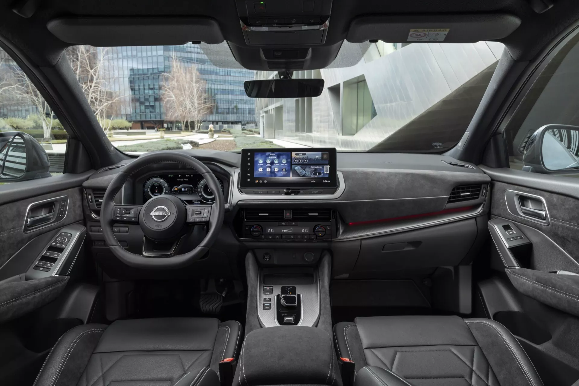Interiér modelu Nissan Qashqai 2024 (J12-Facelift) zobrazující přístrojovou desku, přední sedadla a městské prostředí přes čelní sklo. Vlastnosti zahrnují výrazný volant s ovládacími prvky, digitální infotainment systém, ovládání klimatizace, automatickou převodovku, držáky na nápoje, úložné přihrádky, sportovní sedadla podobná kůži s kontrastním šitím a ambientní osvětlení interiéru.