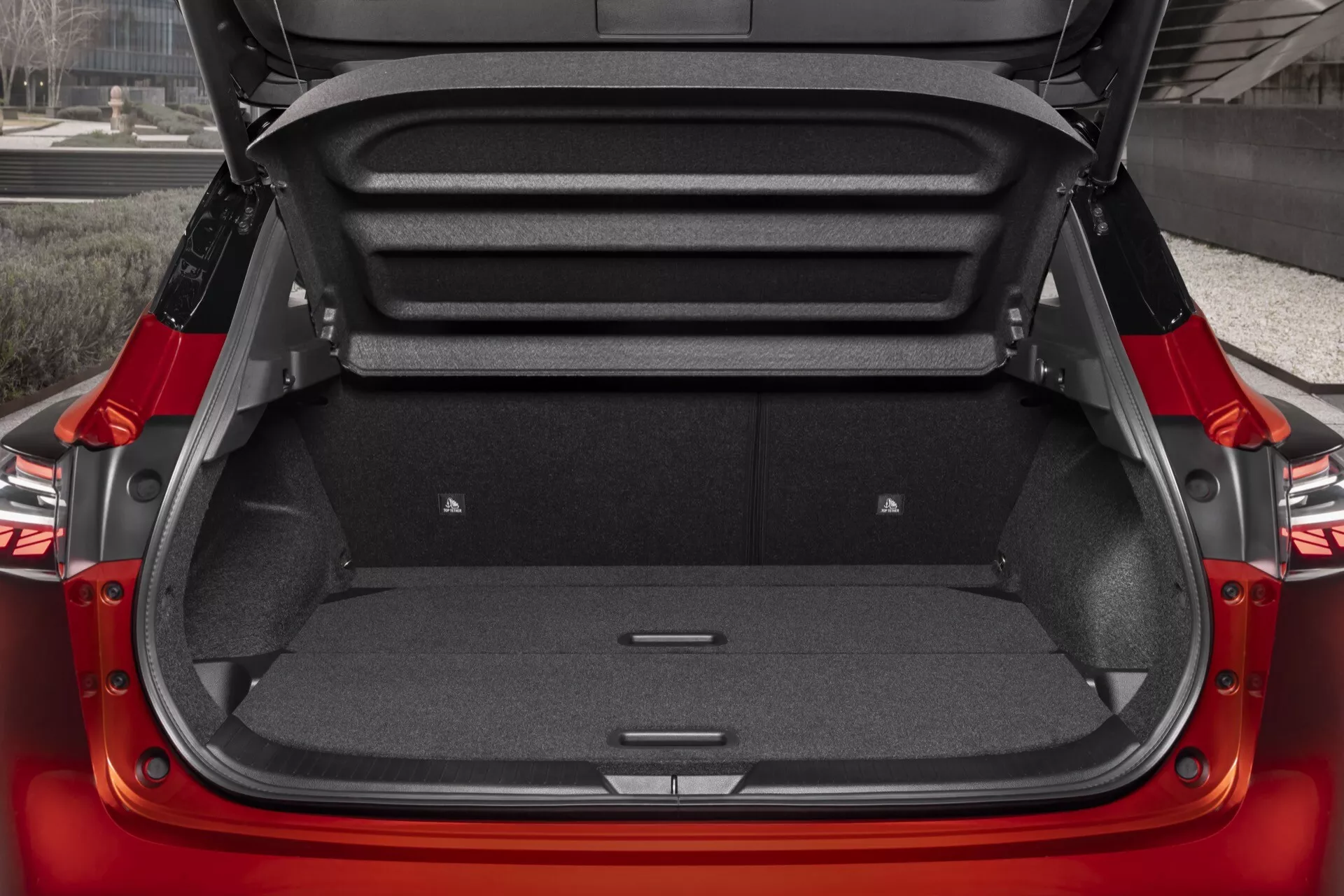 Detailní pohled z vysokého úhlu na zavazadlový prostor vozidla Nissan Qashqai 2024 (facelift J12). Otevřený kufr s čistým, elegantním designem, červenou exteriérem a rovnou tmavou kobercovou podlahou a bočními stěnami s přihrádkami pro nouzové vybavení. Zadní sedadla mají 60/40 skládací design k rozšíření nákladového prostoru.