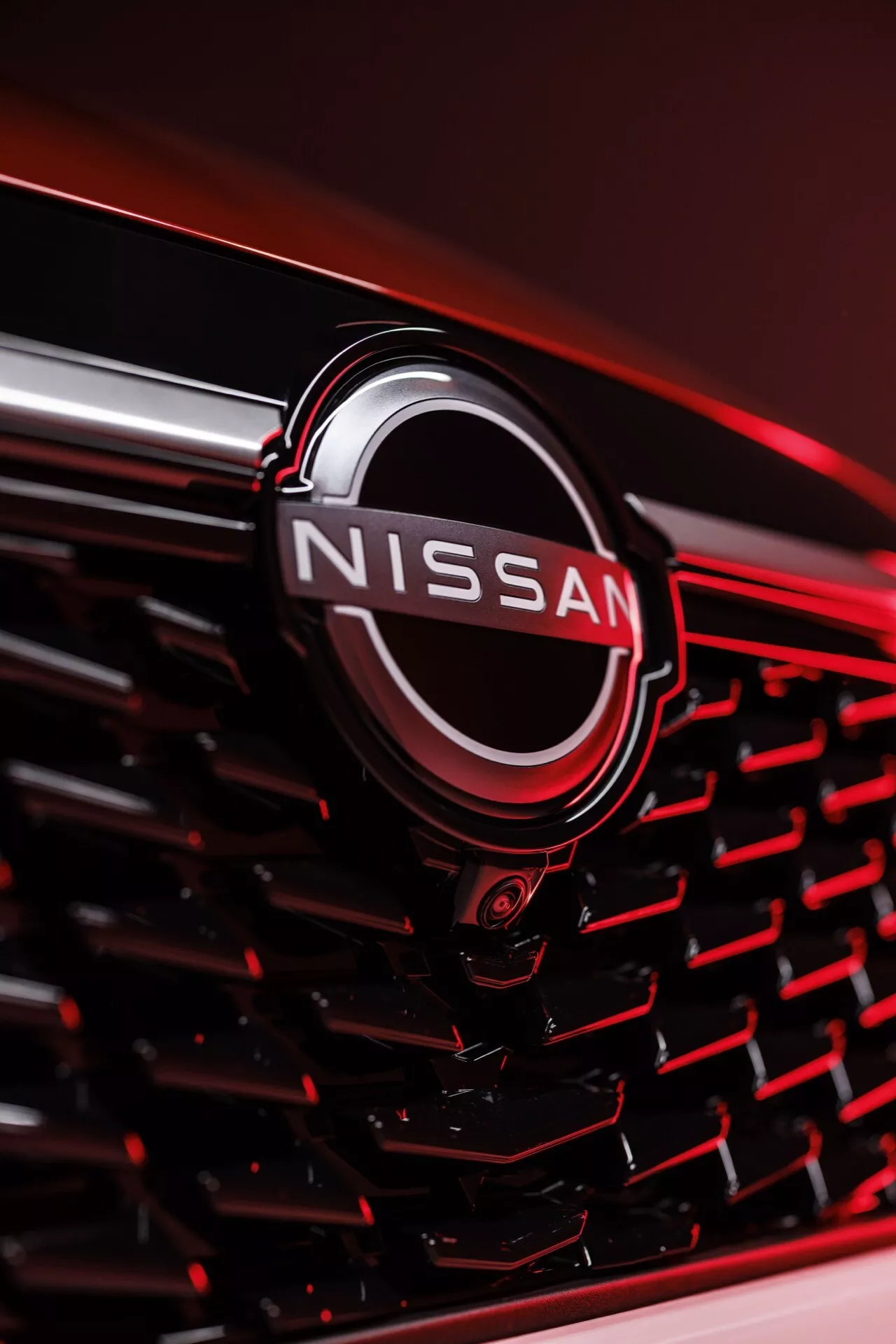 Blízký pohled na přední mřížku 2024 Nissan Qashqai (J12-Facelift) v červené barvě, zdůrazňující chromované logo Nissan s černým výplní. Emblém je uzavřen v kruhu, ústředním prvkem designu mřížky s černým šestiúhelníkovým vzorem. Fotografie je pořízena z nakloněné, pravé perspektivy s malou hloubkou ostrosti, která se ostře zaměřuje na emblém, zatímco okraje se postupně rozmazávají, zachycující odrazy v chromových prvcích.