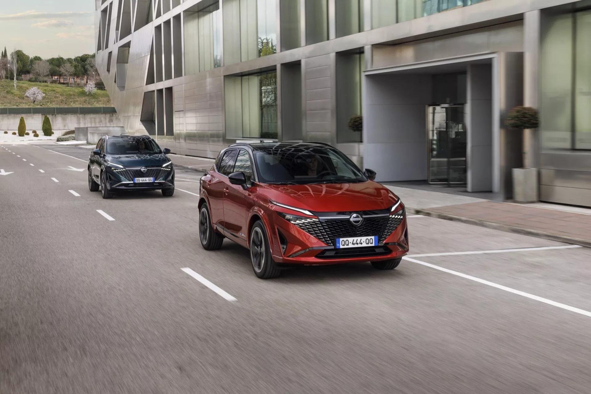 Dva vozy Nissan Qashqai 2024 (J12-Facelift) jedou po městské ulici za denního světla. Přední vůz je červený hatchback zobrazený z tříčtvrtečního čelního pohledu, který zdůrazňuje jeho moderní design s ostrými liniemi, aerodynamický profil, hranatá LED světla, aerodynamická zpětná zrcátka a viditelné senzory v masce, které naznačují pokročilé asistenční systémy řidiče. Zadní vůz, možná černý nebo tmavě modrý, je podobný model zachycený ze zadního tříčtvrtečního pohledu, viditelné jsou zadní světla a zadní okno. Oba vozy jsou v pohybu na víceproudé silnici s přerušovanými pruhy a moderním městským architektonickým prostředím.