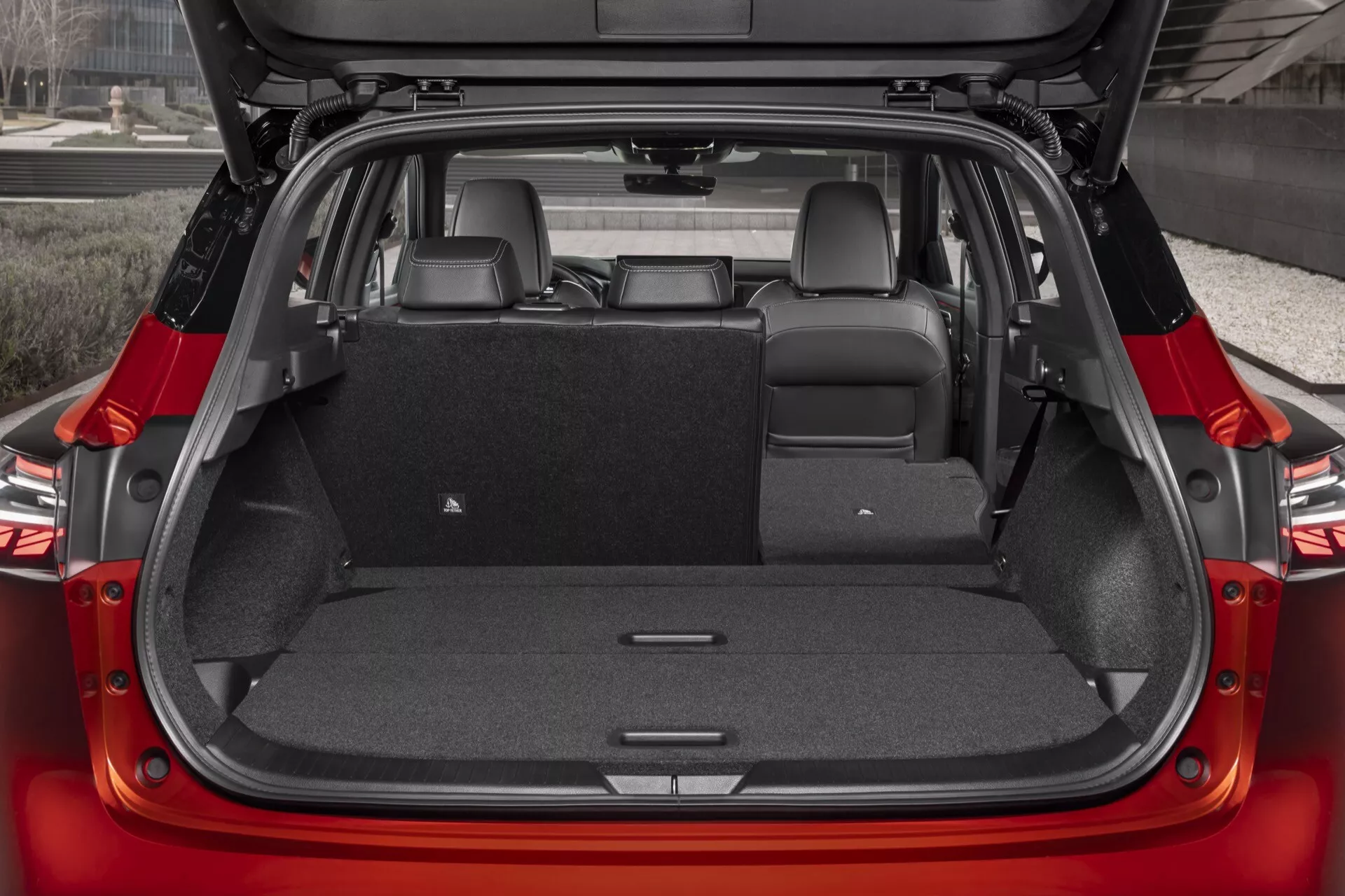 Obrázek ukazuje zadní interiér vozu Nissan Qashqai 2024 (J12-facelift) z mírně zvýšené perspektivy zezadu. Zadní dveře jsou otevřené a odhalují čalouněný prostor kufru s částečně sklopeným zadním sedadlem, čímž se ukazuje jeho rozdělená sklápěcí funkce, která rozšiřuje úložný prostor. Chybí zde háky na náklad, síťky a napájecí zásuvky. Koberec tmavé šedé nebo černé barvy je praktický na zakrytí nečistot. Na pravé straně je světlo interiéru, které osvětluje prostor kufru. Městské prostředí na pozadí je rozmazané, což zdůrazňuje interiér vozu.