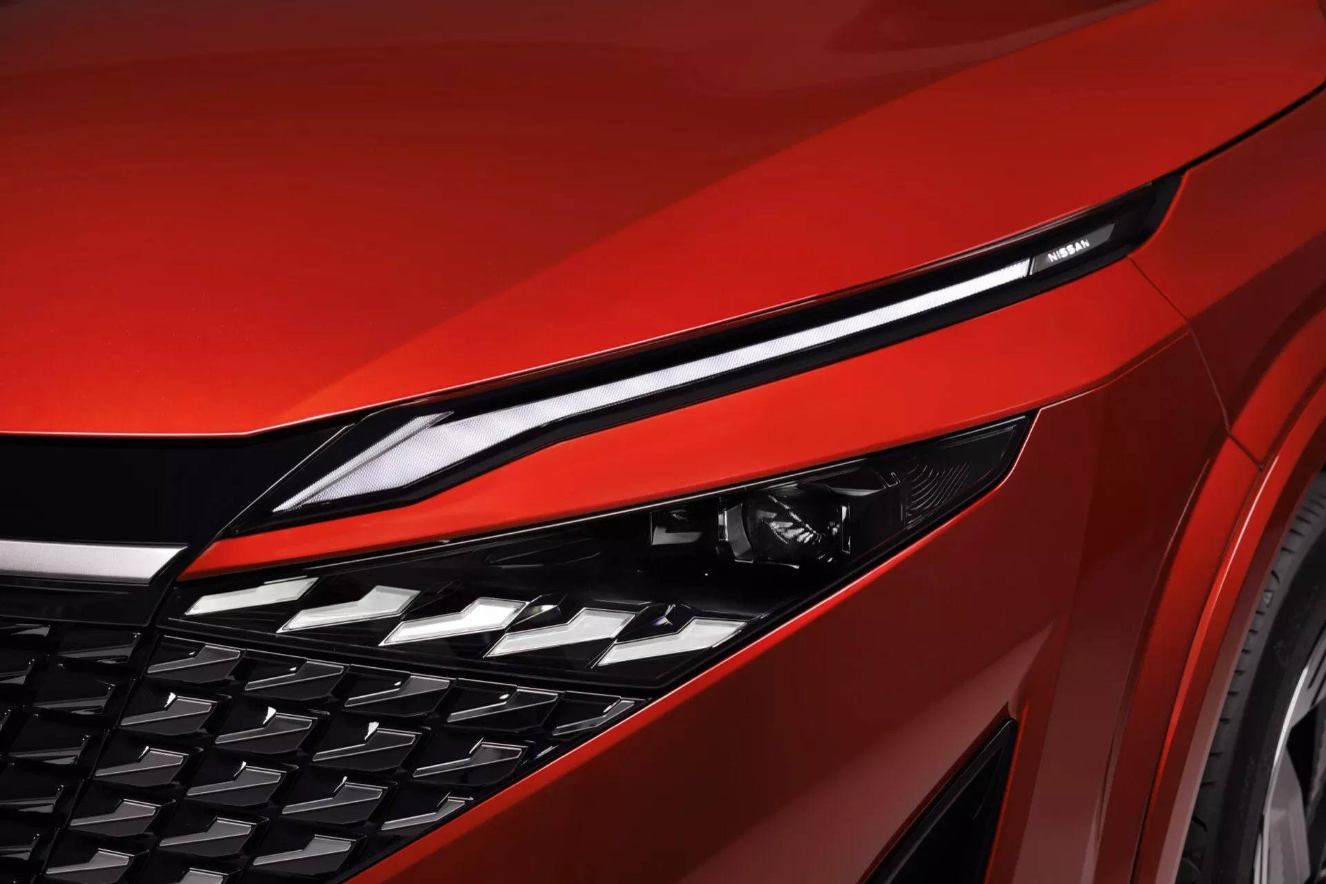 Detailní pohled na přední část Nissan Qashqai 2024 (J12-facelift), zdůrazňující elegantní a protáhlý LED světlomet, výrazný DRL pás, velkou hranatou mřížku a částečné pohledy na kapotu a kolo. Odvážná červená barva a záběr z nízkého úhlu zdůrazňuje agresivní design, pokročilé osvětlení, aerodynamický design mřížky a sportovní kontury kapoty, což ukazuje na kvalitní řemeslné zpracování a moderní technologie.