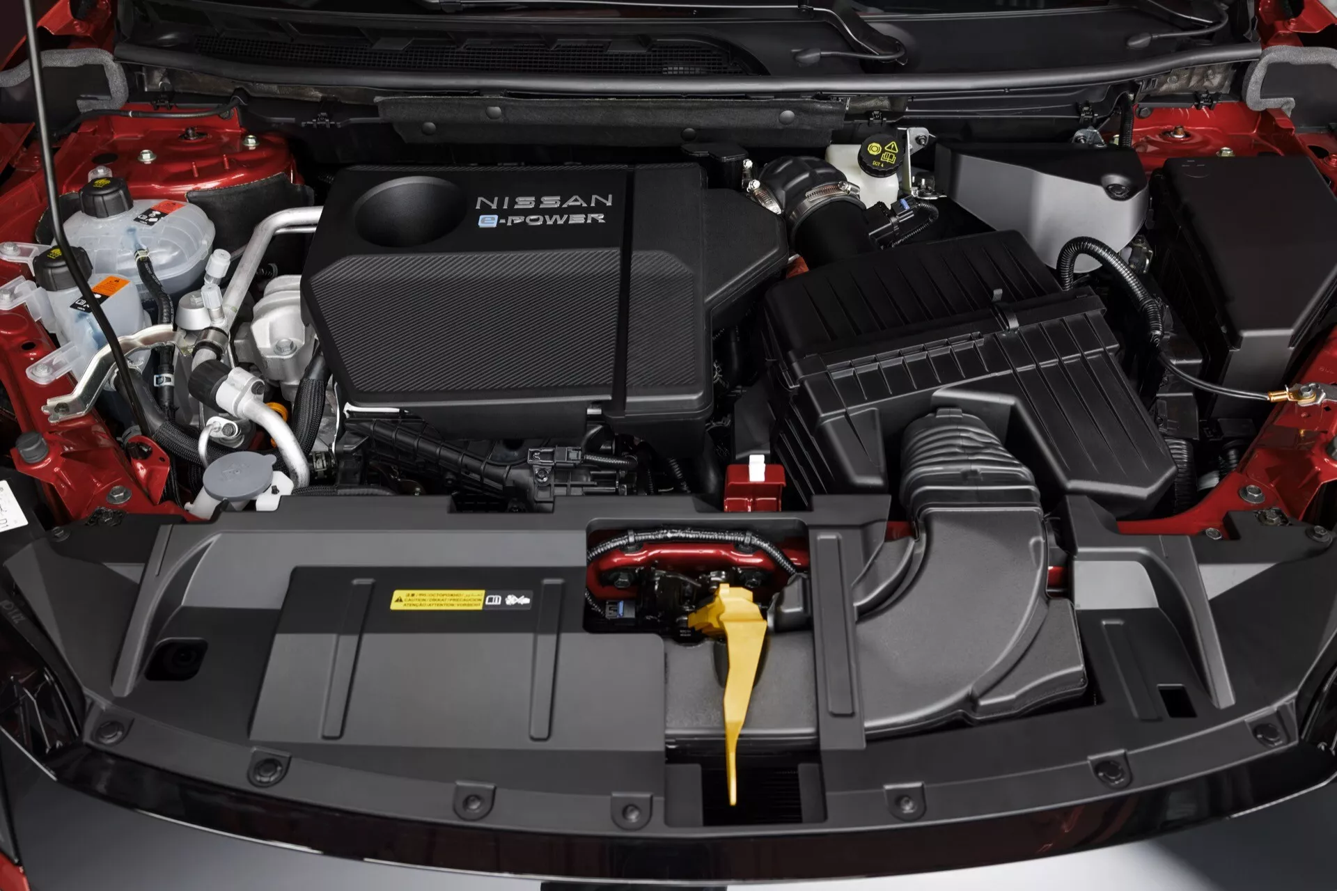 Detailní pohled shora na motorový prostor Nissan Qashqai 2024 (J12-Facelift) s otevřenou kapotou, uprostřed se značkovým plastovým krytem motoru 'NISSAN'. Viditelné komponenty zahrnují na pravé straně systém sání vzduchu, vlevo žlutý měrku oleje a na horní levé straně nádržku brzdové kapaliny se žlutým víčkem. Různé nádoby na kapaliny, kabeláž a potrubí jsou pečlivě uspořádány a zvýrazňují pokročilé automobilové technologie moderního vozidla.