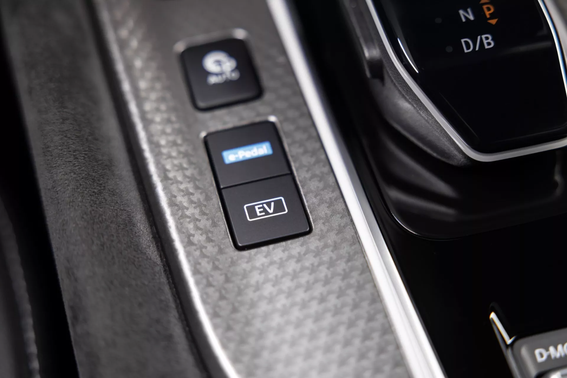 Detailní záběr interiéru vozu Nissan Qashqai 2024 (J12-Facelift) se zaměřením na tlačítka EV režimu a asistenta parkování. Tlačítka jsou umístěna na texturovaném panelu připomínajícím uhlíková vlákna s kartáčovaným kovovým lemem, zdůrazňujícím moderní design a vyšší modelovou třídu. Nad tlačítky je viditelné řadicí páka s označením 'P' pro parkování a další částečně viditelné ovládací prvky. Osvětlení a stíny zvýrazňují ergonomické a estetické detaily.