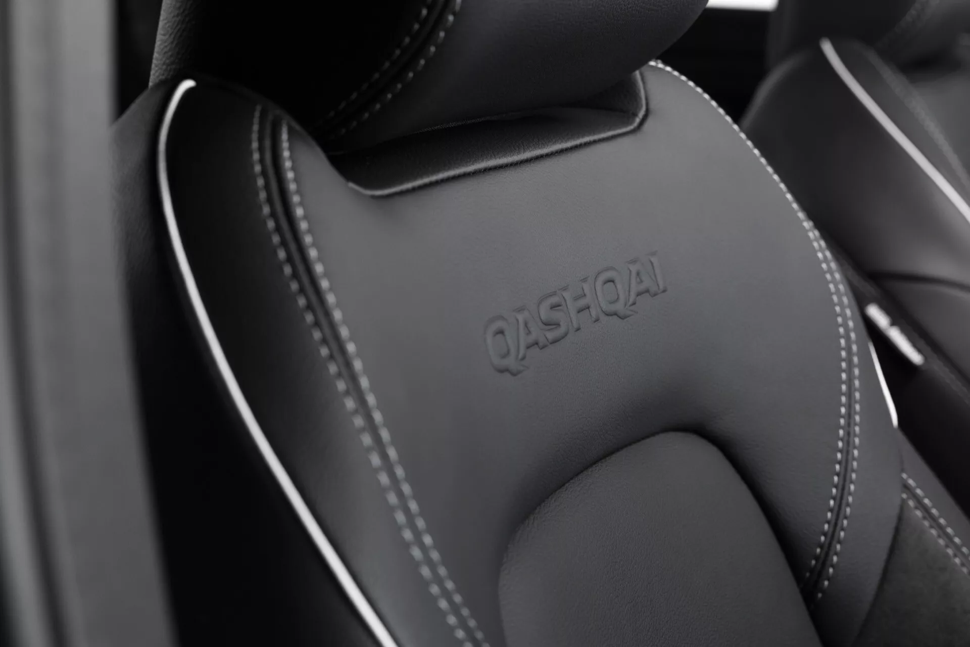 Detailní záběr na interiér Nissan Qashqai 2024 (J12-Facelift) se zaměřením na horní část sedadla řidiče nebo spolujezdce. Opěrka hlavy výrazně zobrazuje vyražený název 'QASHQAI', s detailním prošitím po švech. Prémiový materiál sedadla, který může být z kůže nebo kvalitní syntetiky, ukazuje monochromatické barevné schéma, převážně v odstínech černé nebo tmavě šedé. Design zdůrazňuje luxusní a odolný přístup. Mělká hloubka ostrosti zdůrazňuje text 'QASHQAI', s jemně rozmazaným popředím a pozadím.