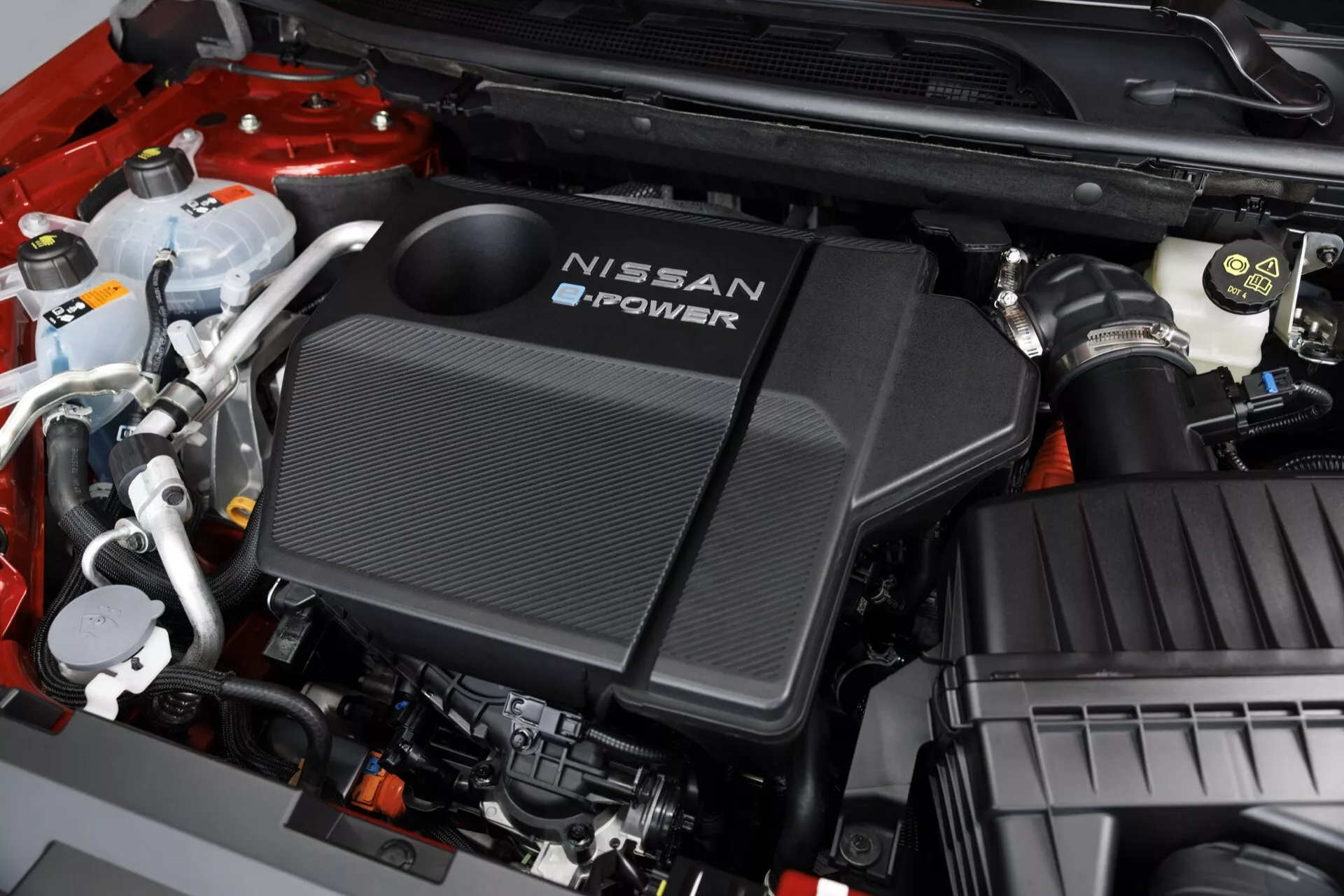 Detailní pohled na motorový prostor Nissan Qashqai 2024 (J12-facelift) z vysokého úhlu zaměřený na kryt motoru. Kryt s nápisem 'NISSAN e-POWER' naznačuje hybridní nebo elektrický pohonný systém. Obsahuje texturu podobnou uhlíkovým vláknům, různé komponenty jako chladicí nádrže a sací potrubí vzduchu a organizovaná elektrická připojení. Barevné schéma zahrnuje černou, bílou, stříbrnou, červenou a žluté krytky. Na pravé straně se nachází krabice vzduchového filtru, která naznačuje pokročilý a dobře udržovaný motorový prostor.
